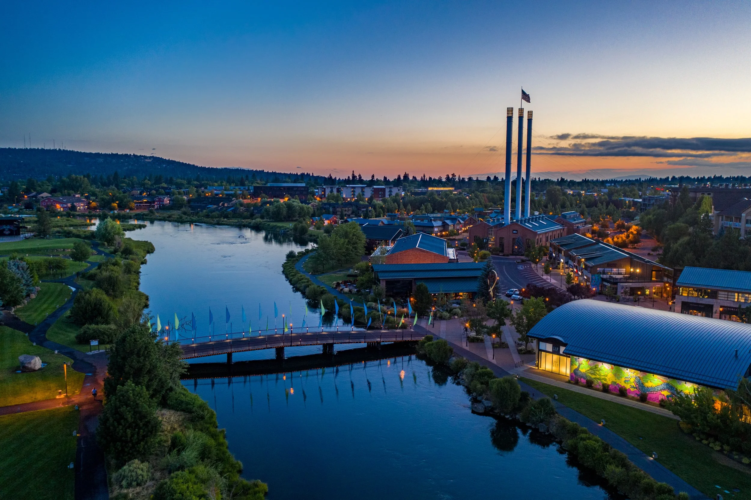 A scenic view of a town at sunset with a river, bridge, and buildings, including three tall smokestacks with an American flag on top.