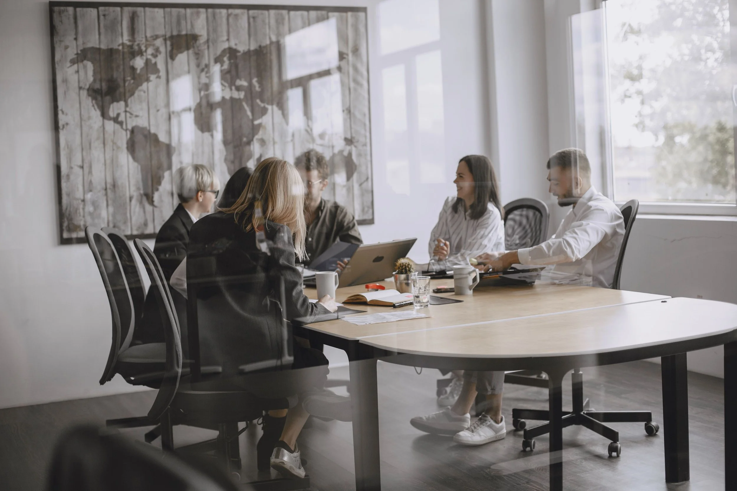 A group of five diverse professionals having a discussion in a modern conference room with large windows, a world map art piece on the wall, and a wooden table with laptops, notebooks, coffee cups, and water glasses.