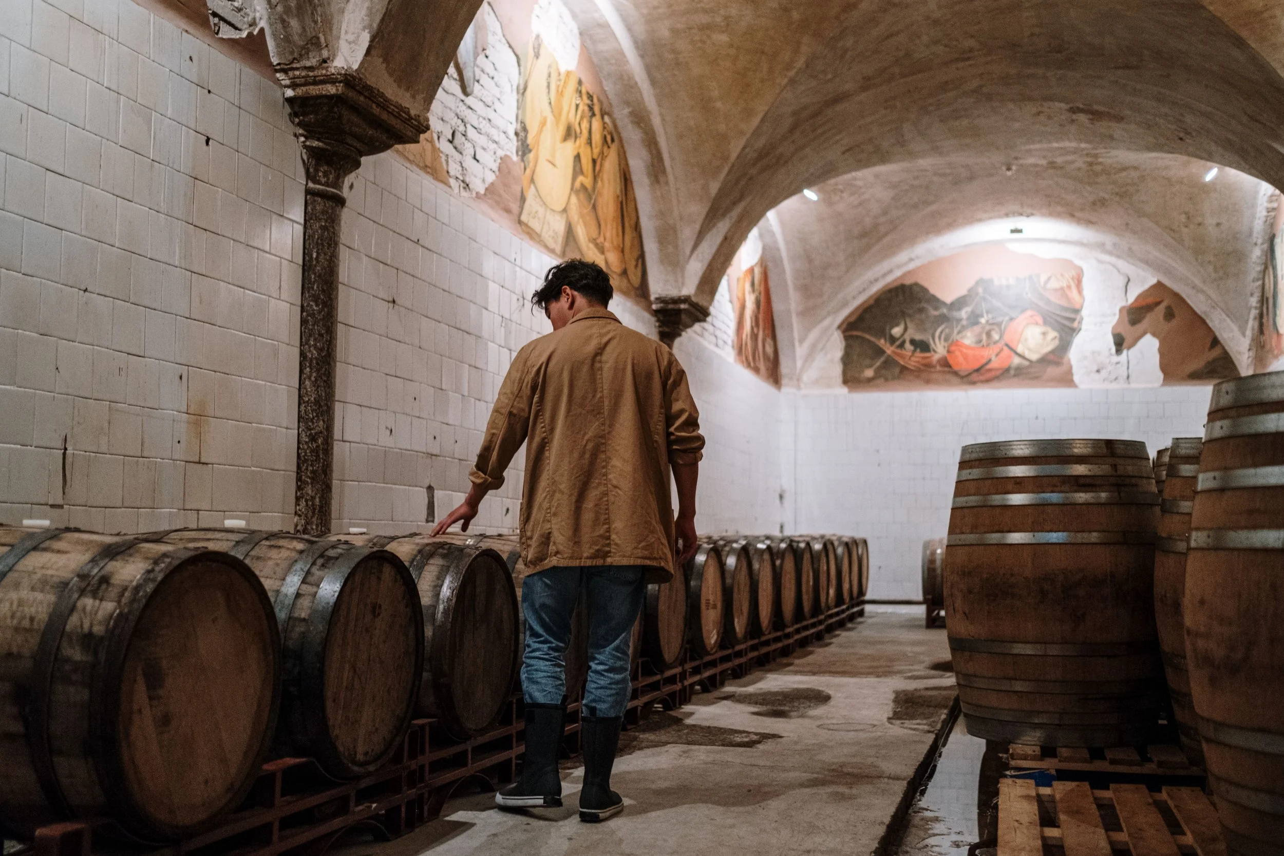 A man in a brown jacket standing in a wine cellar with wooden barrels and frescoed arch ceilings.