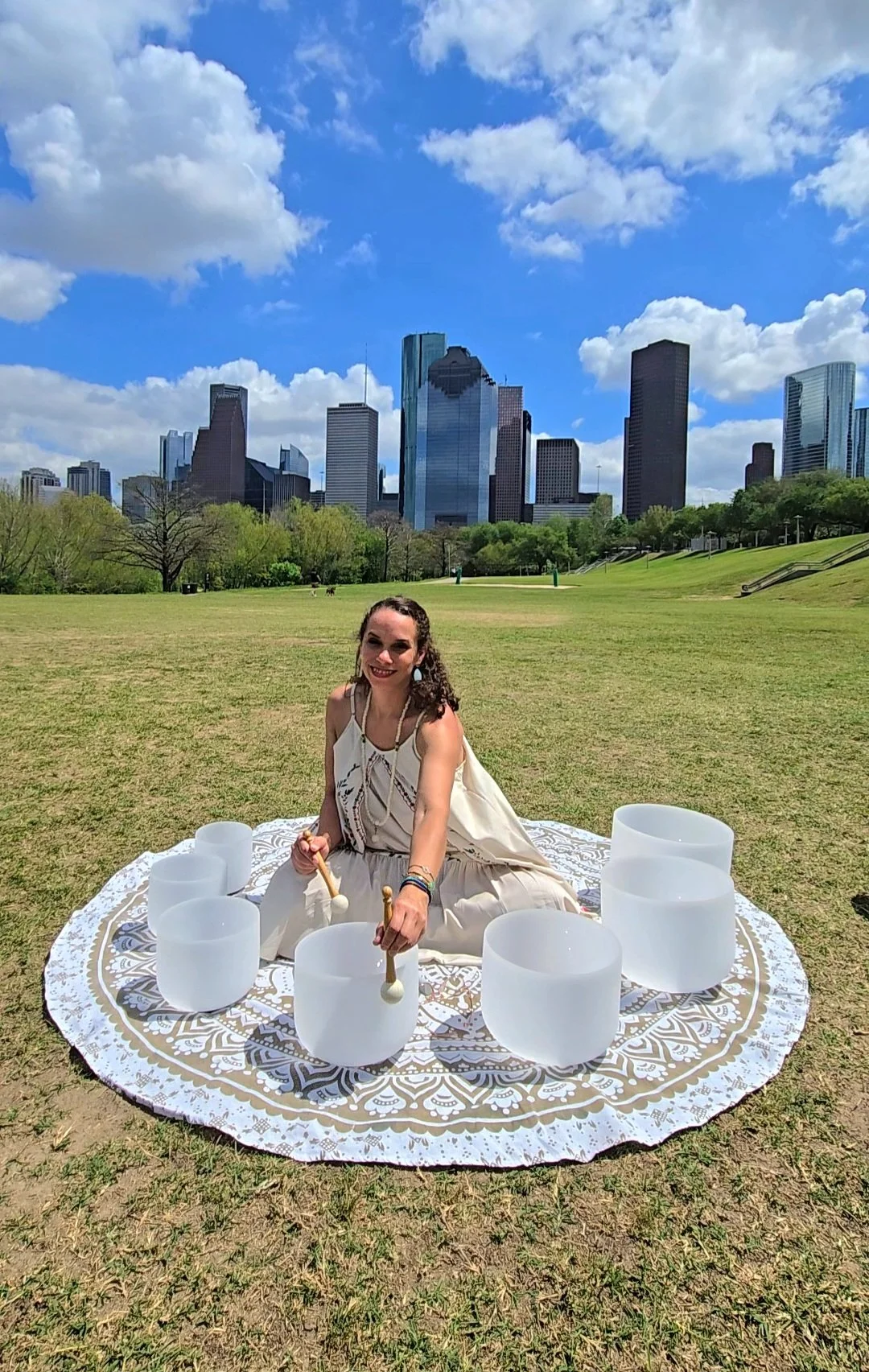 A woman sitting on a patterned cloth in a grassy park with a city skyline in the background. She is smiling and playing crystal singing bowls, with a bright sky and scattered clouds overhead.