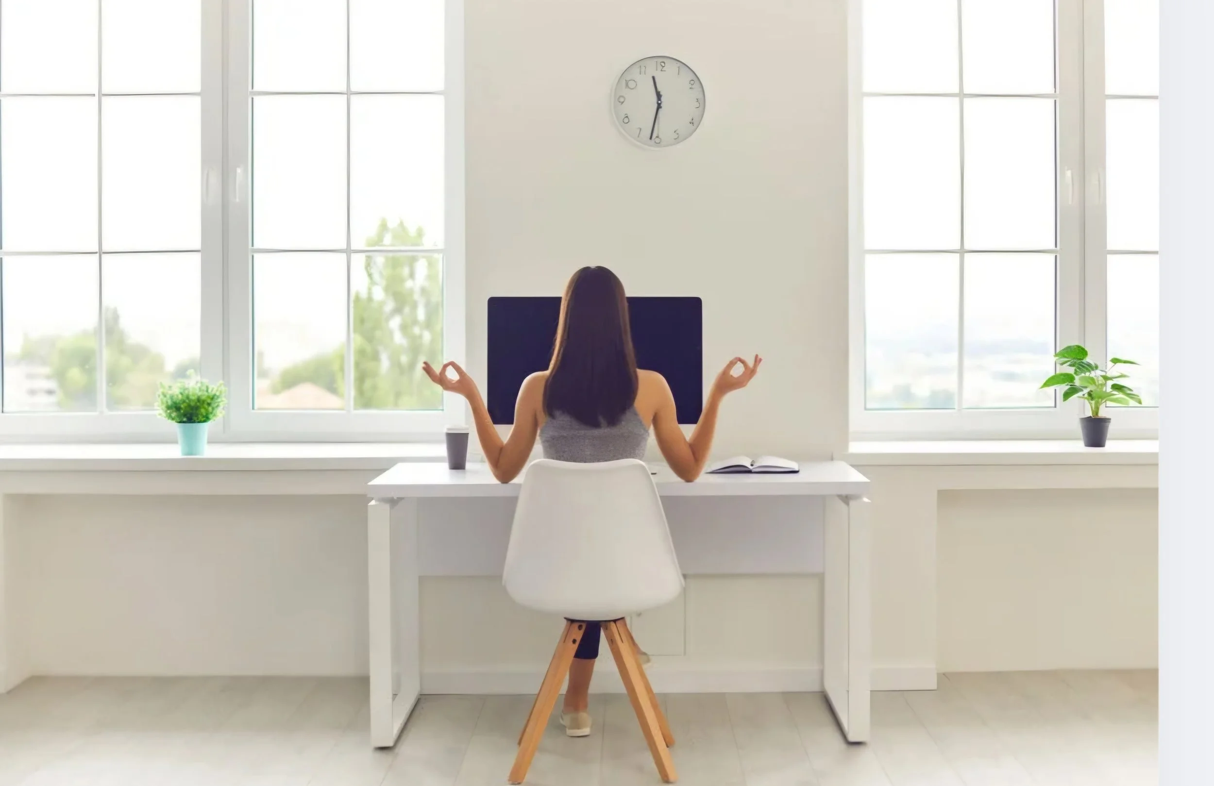 A woman practicing meditation at a white desk in front of large windows with a clock showing 12:01. She is sitting in a white chair, facing away, with her hands in a mudra position.