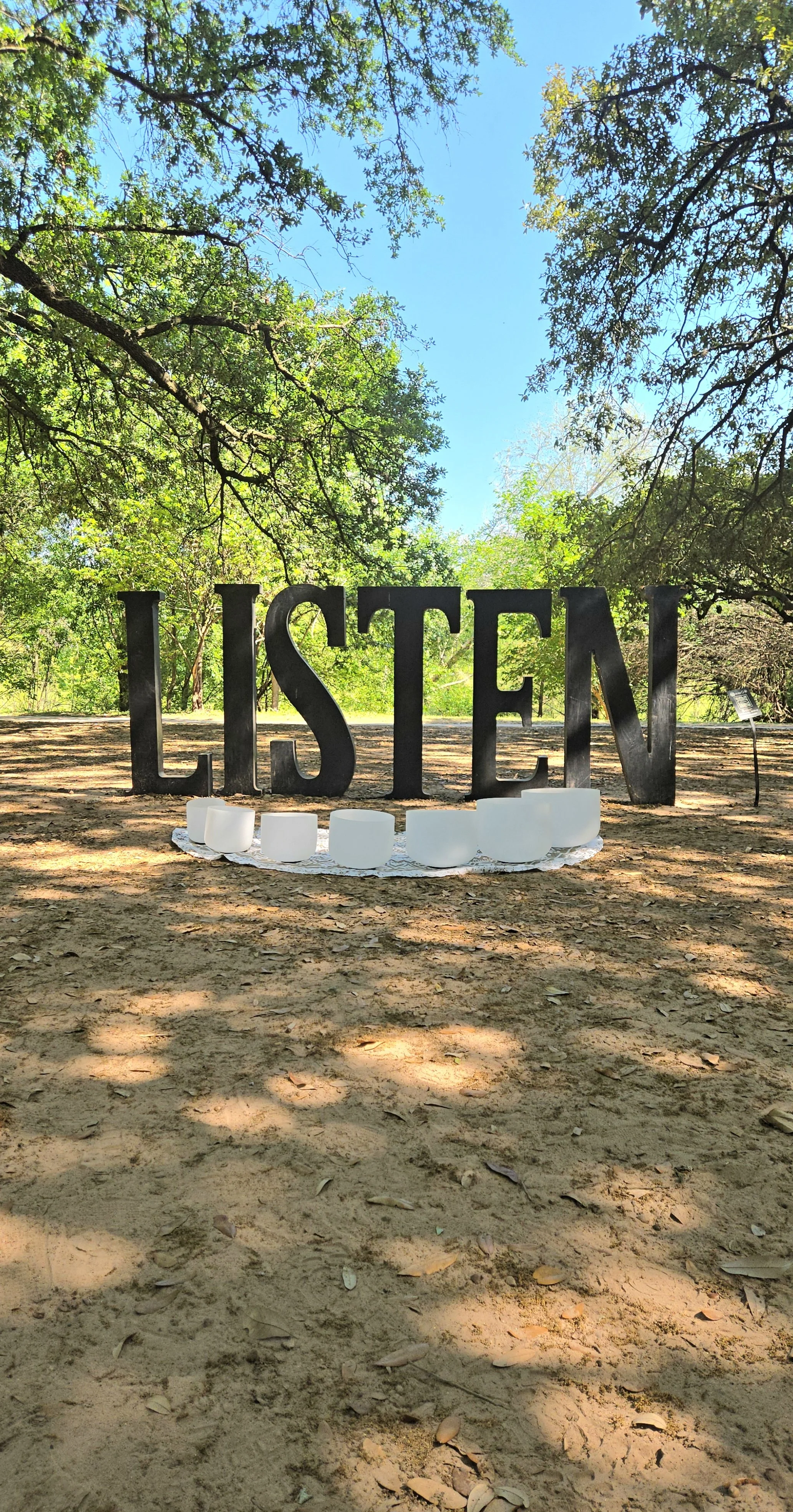 Large black letters spelling 'LISBON' are displayed on the ground in a wooded area with trees and a clear blue sky in the background. White rocks are placed at the base of the letters.
