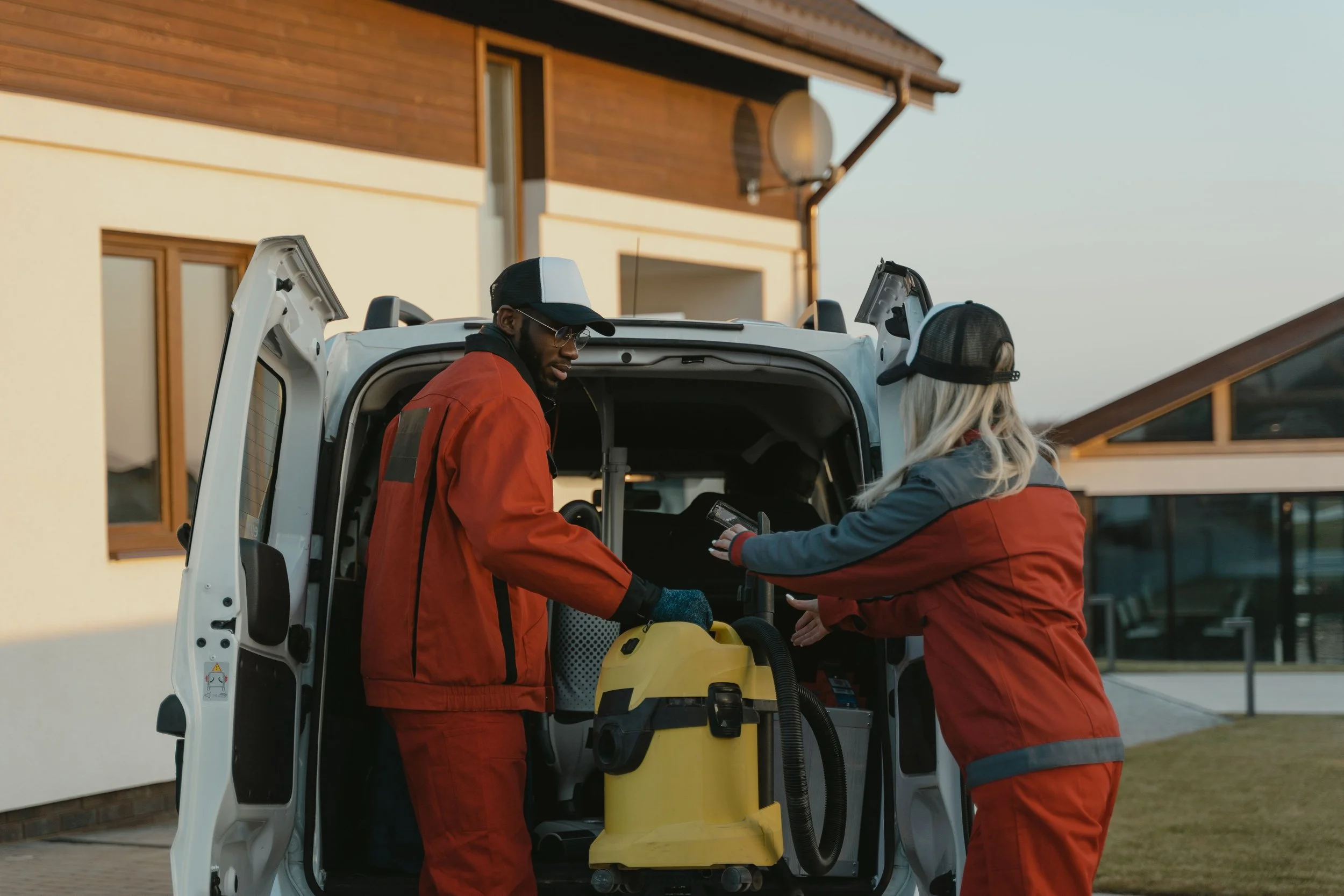 Two emergency responders in red jackets are unloading medical equipment from a white emergency vehicle in front of a modern house with wooden accents during daylight.