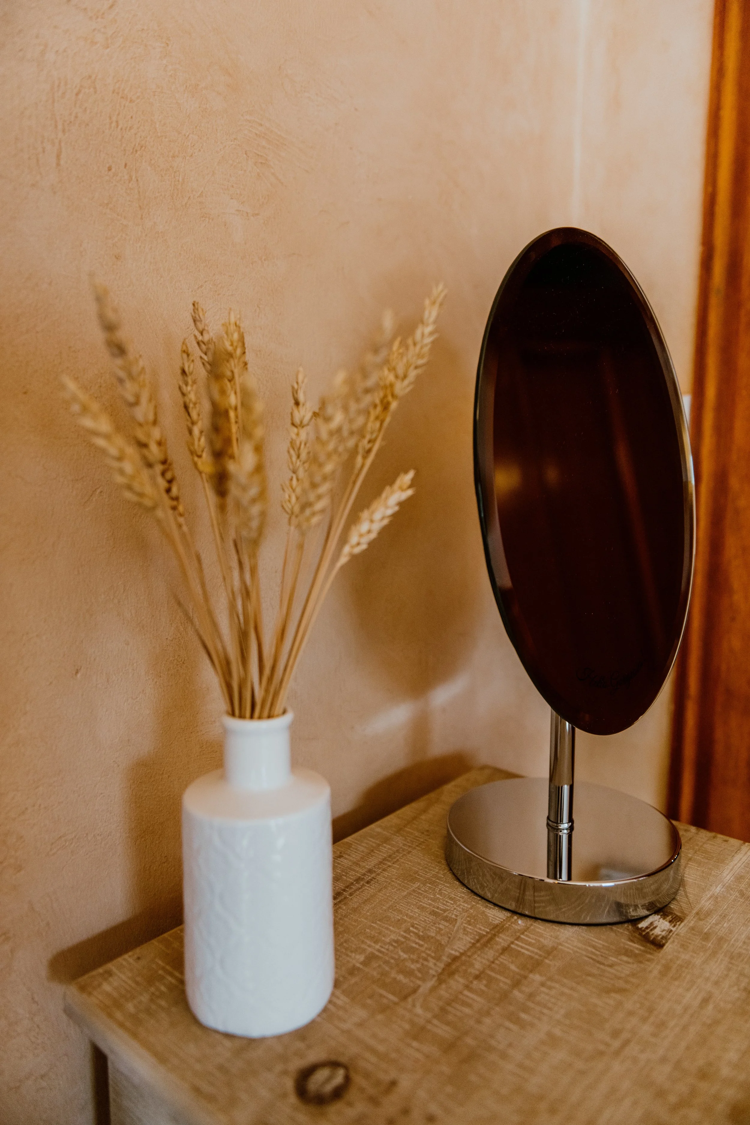 A wooden table with a white vase holding dried wheat stalks and a black oval mirror with a metal stand, against a beige wall.