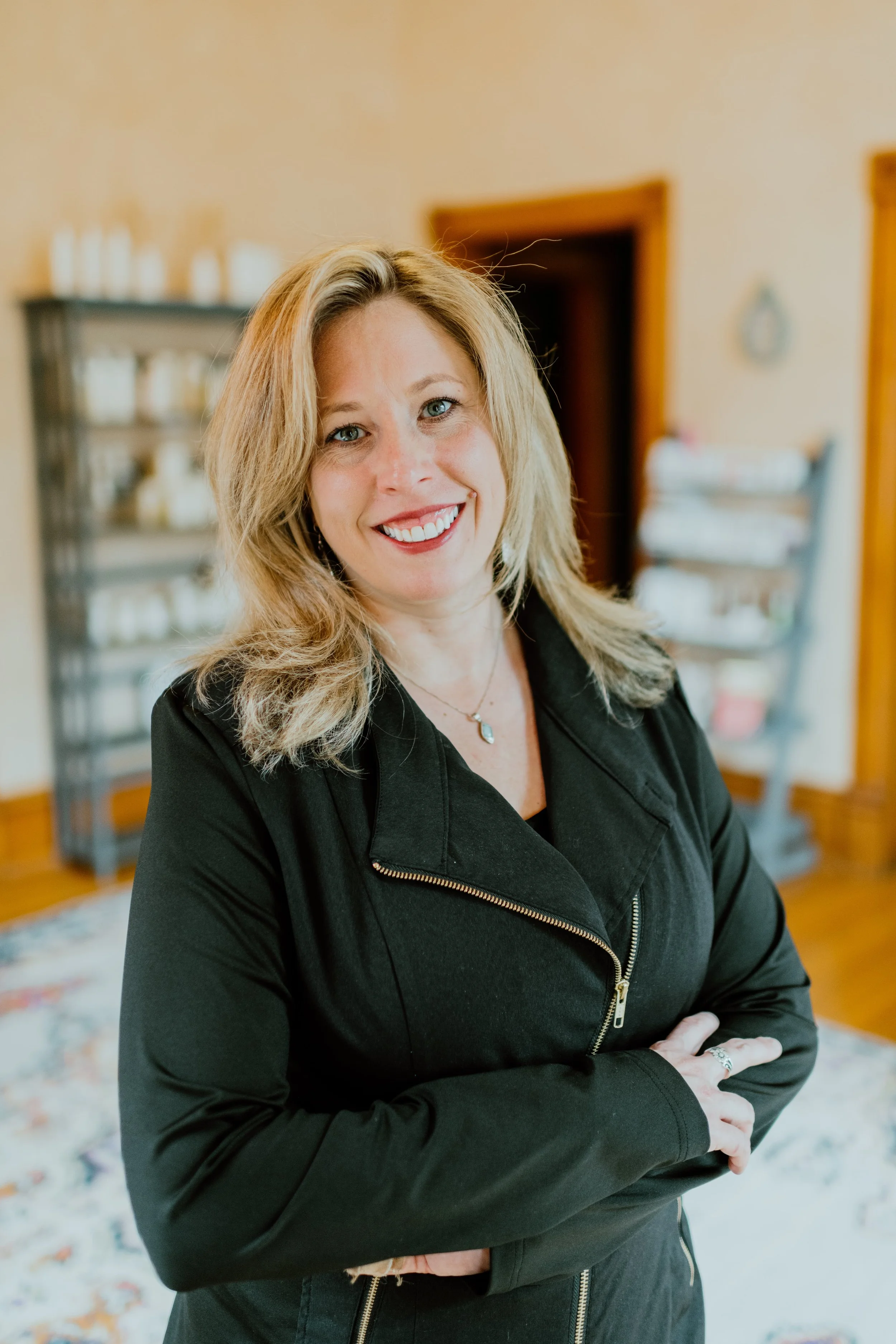 A woman with shoulder-length blonde hair, wearing a black jacket with a zipper, smiling with her arms crossed inside a room with a bookshelf and an open door in the background.