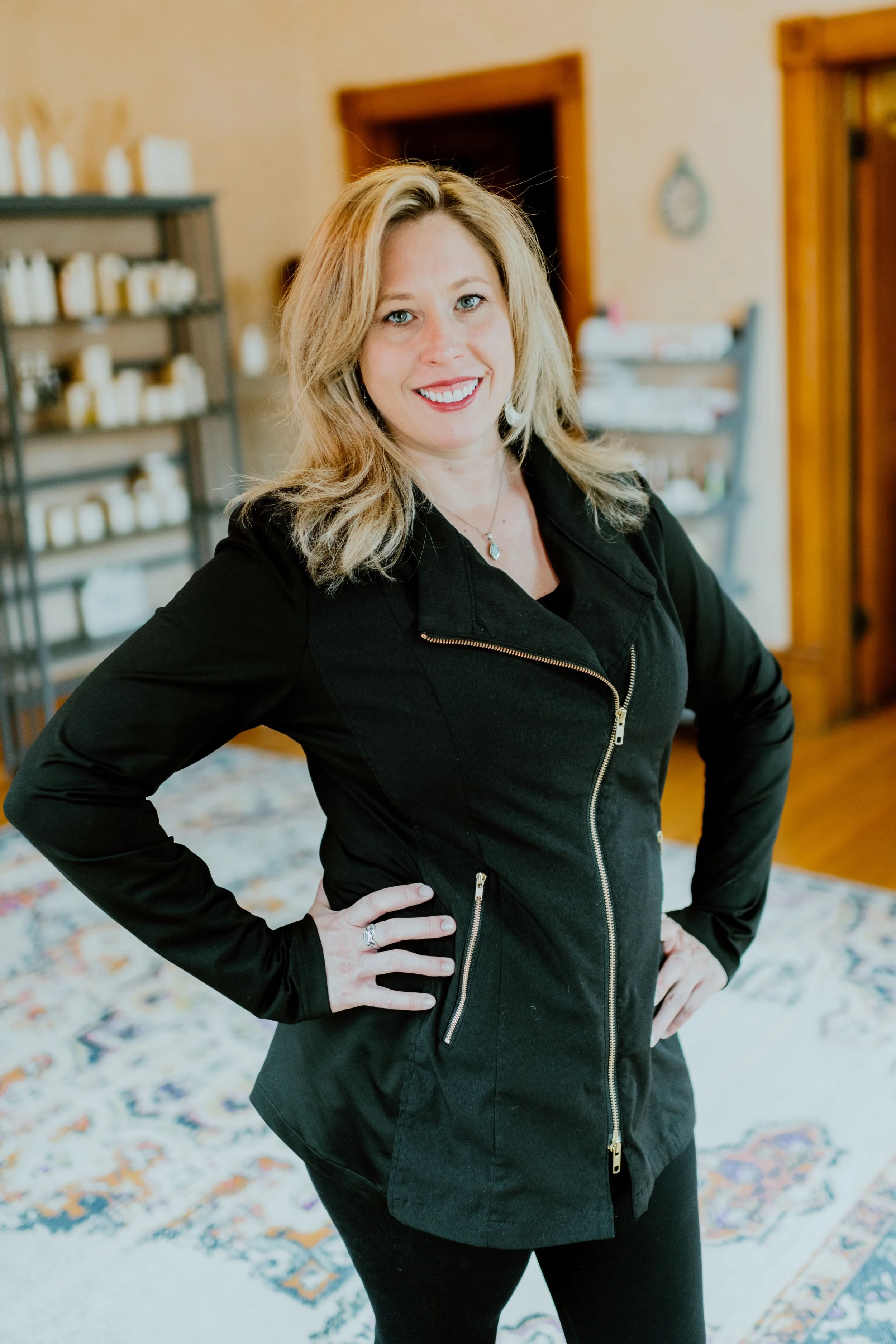A woman with blonde hair wearing a black jacket and black pants, smiling with her hand on her hip, standing in a room with wooden door frames, shelves with jars, and a colorful rug.