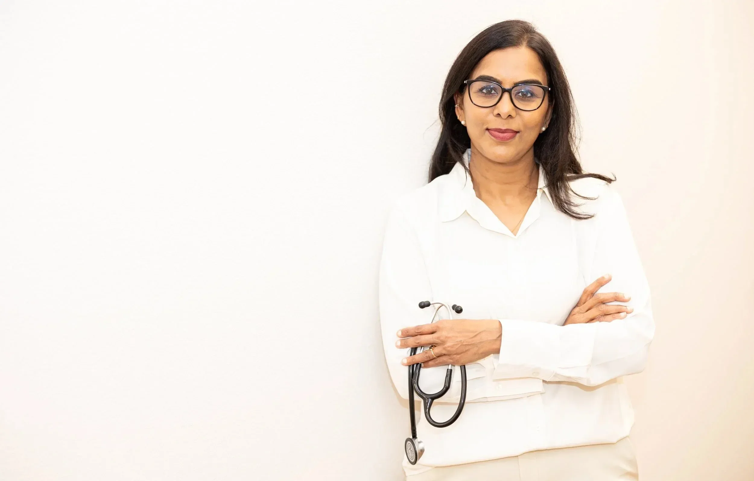 Dr. Maheswari Raja with dark hair and glasses, wearing a white shirt and holding a black stethoscope against a plain white wall background.