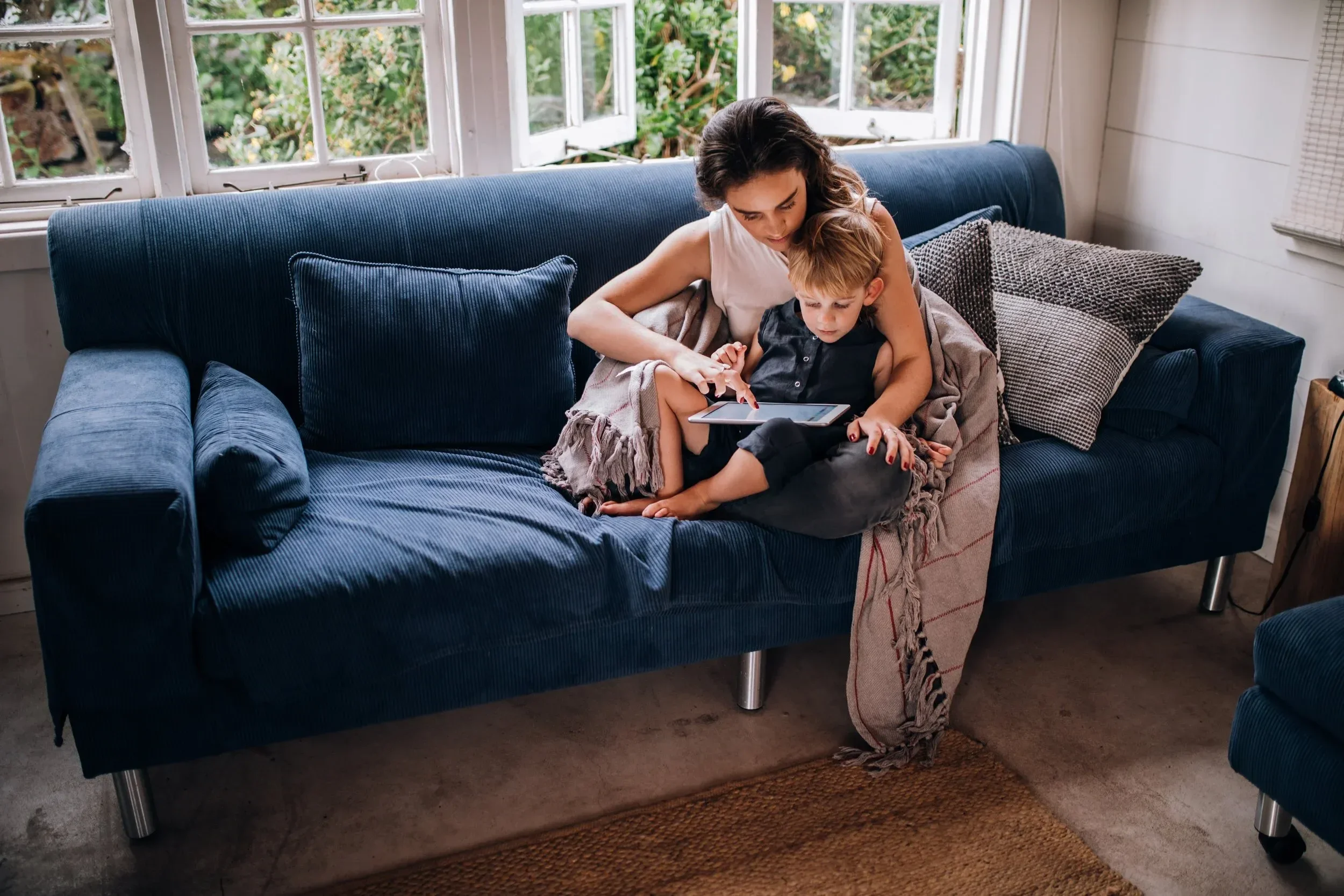 A woman and a young boy sitting on a blue couch by a window. The woman is looking at a tablet, and the boy is sitting on her lap, also looking at the device. There are cushions on the couch and a blanket around their legs. Outside the window, greenery is visible.