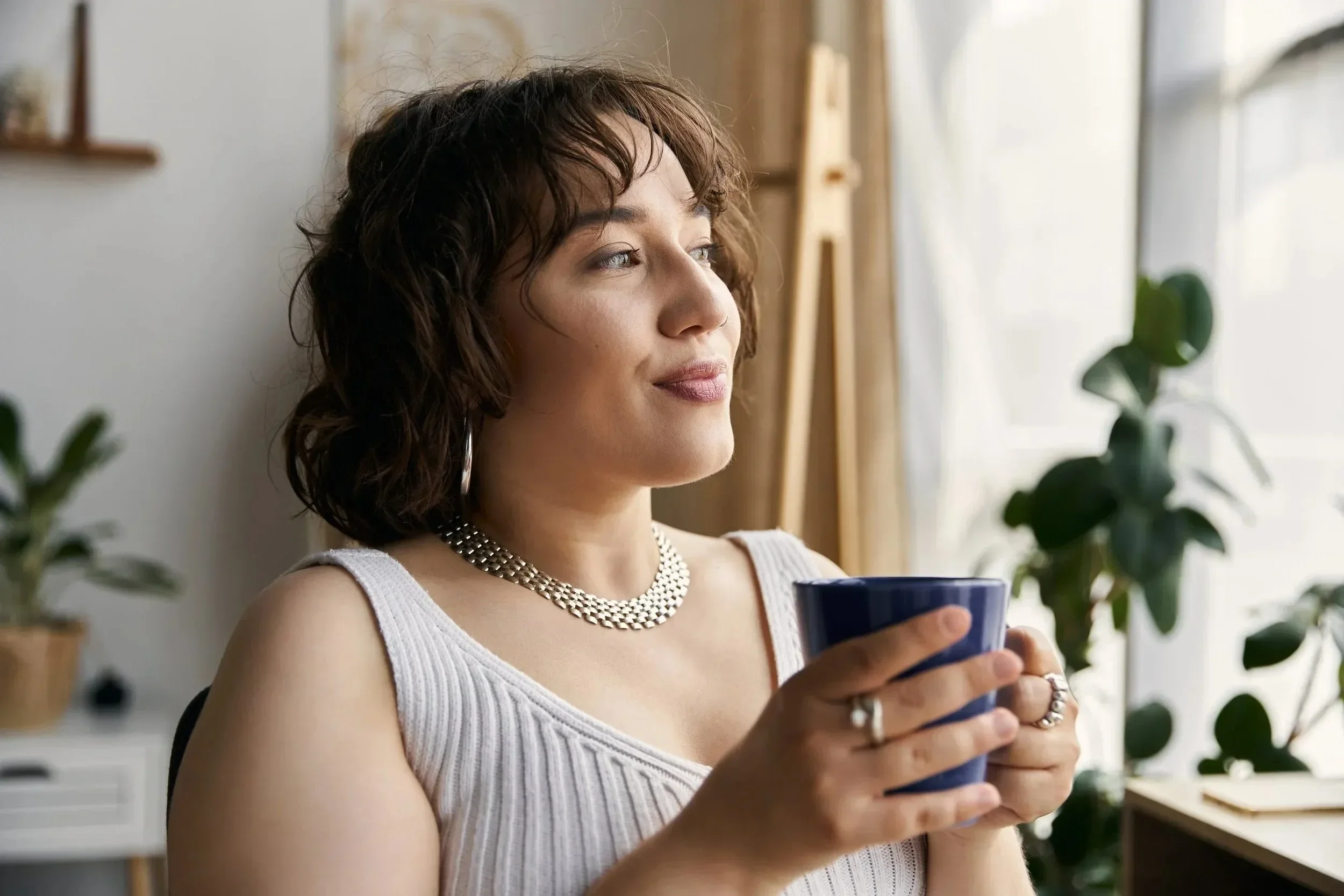 A woman with short, curly brown hair wearing a white tank top and a silver necklace, holding a blue mug and looking out a window in a room decorated with plants.