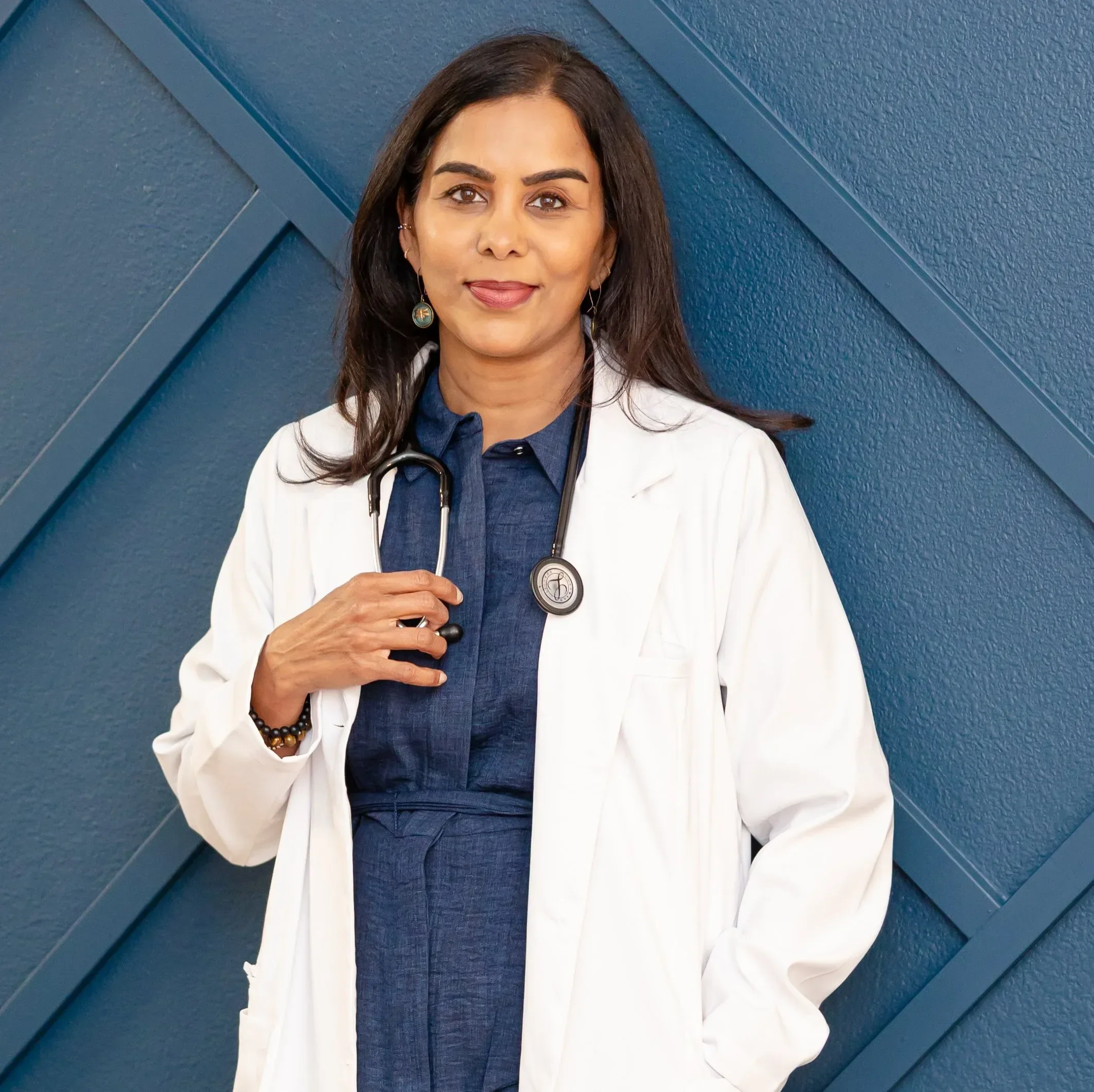 Dr. Maheswari Raja in a white medical coat with a stethoscope around her neck, standing in front of a blue geometric wall.