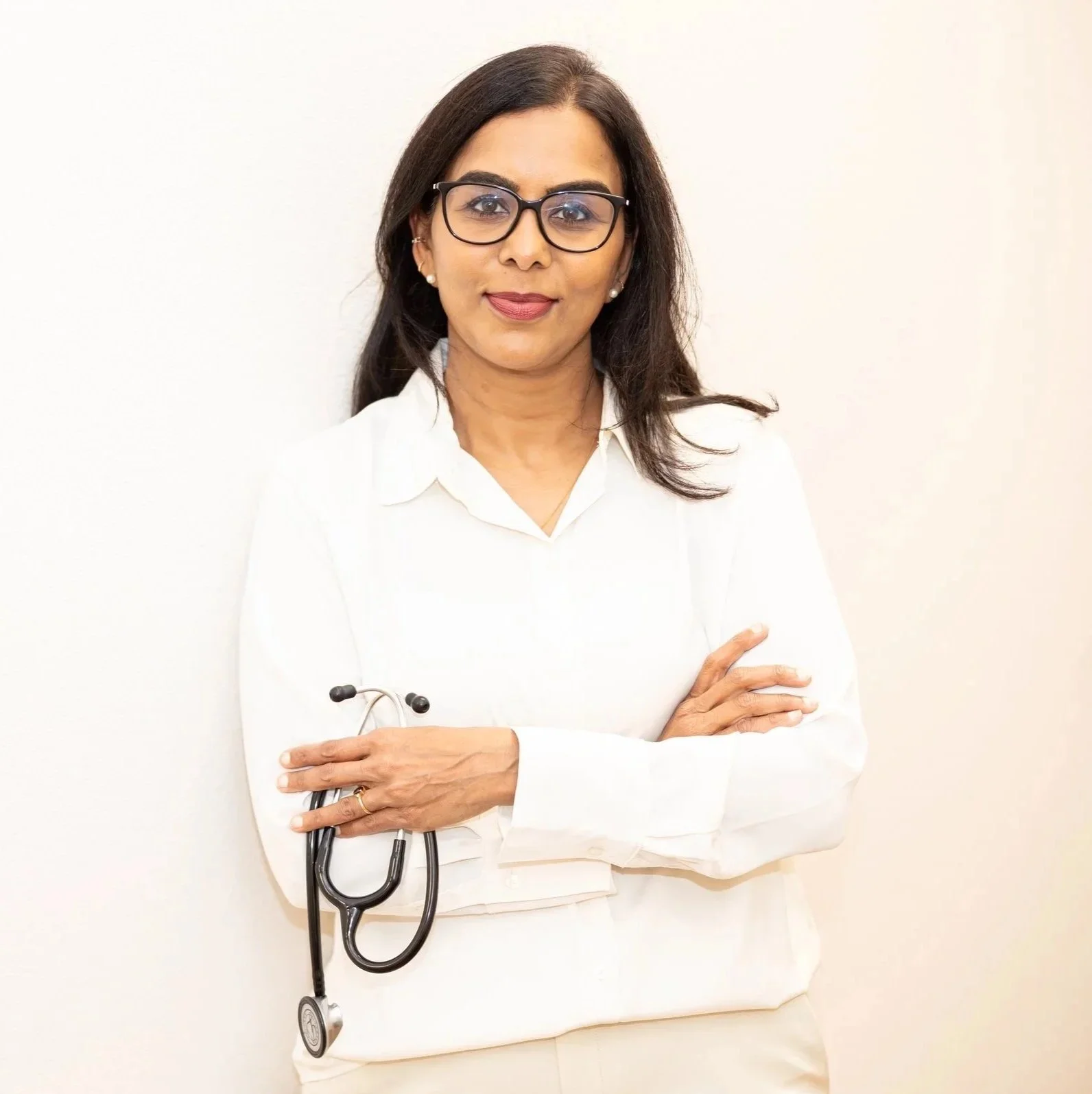 Dr. Maheswari Raja in glasses and a white blouse standing against a plain wall, holding a stethoscope in her hand, with arms crossed.