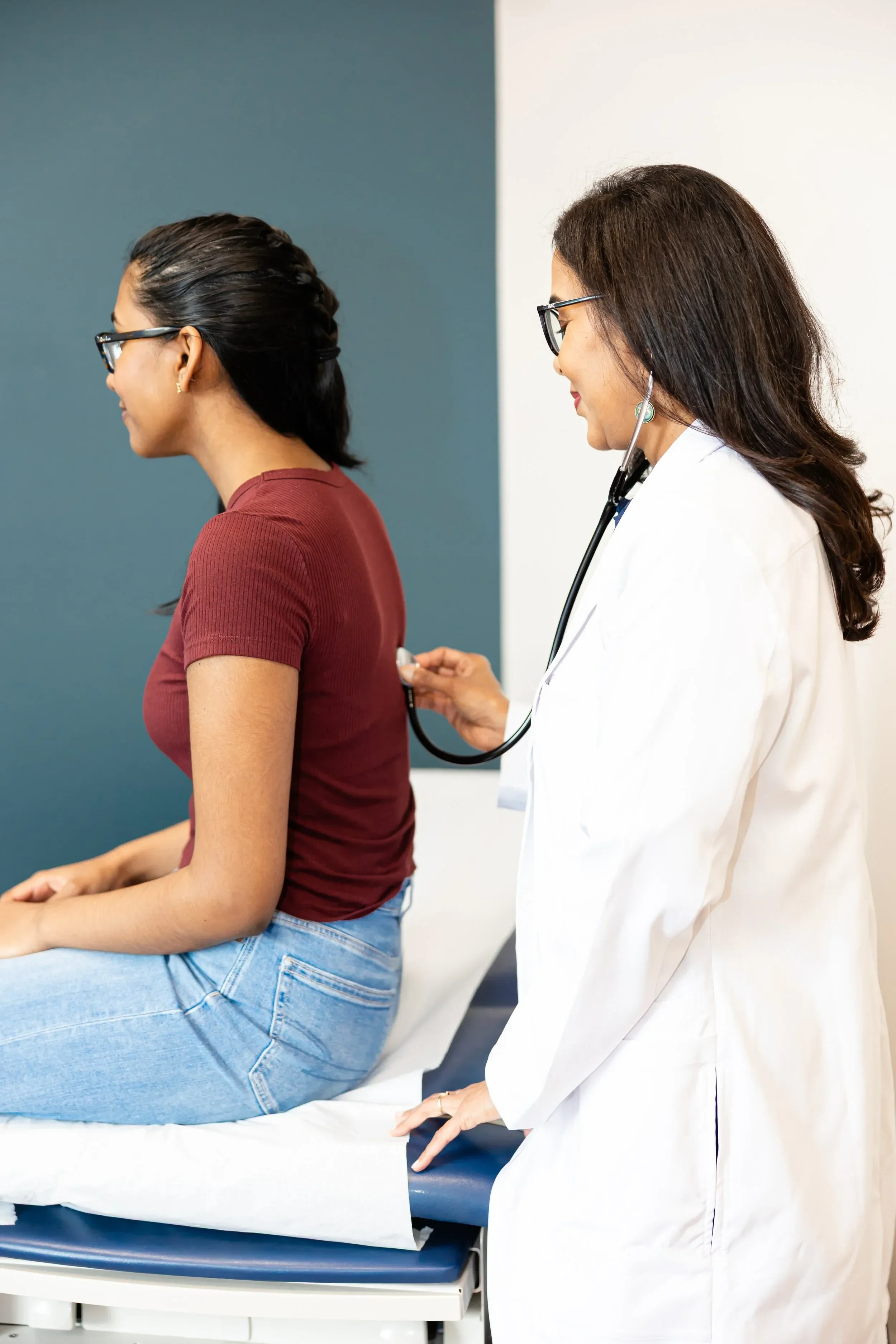 Dr. Maheswari Raja is checking a  woman sitting on an examination table, wearing a maroon shirt and jeans, with her hands on her lap. The background features a two-tone wall, dark blue on one side and white on the other.