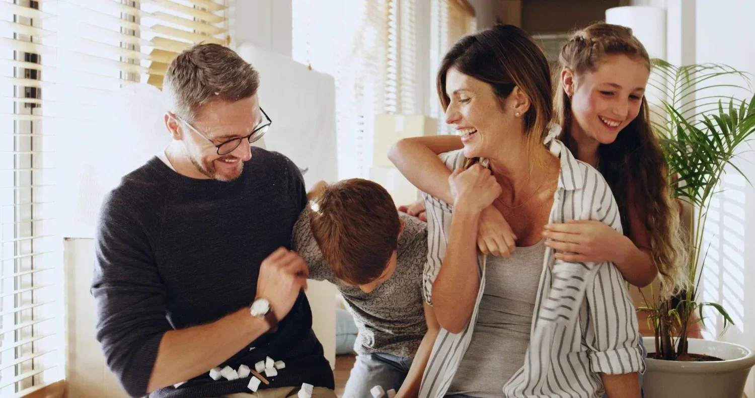 A family laughing and playing together inside a bright living room, engaging in a playful pillow fight.