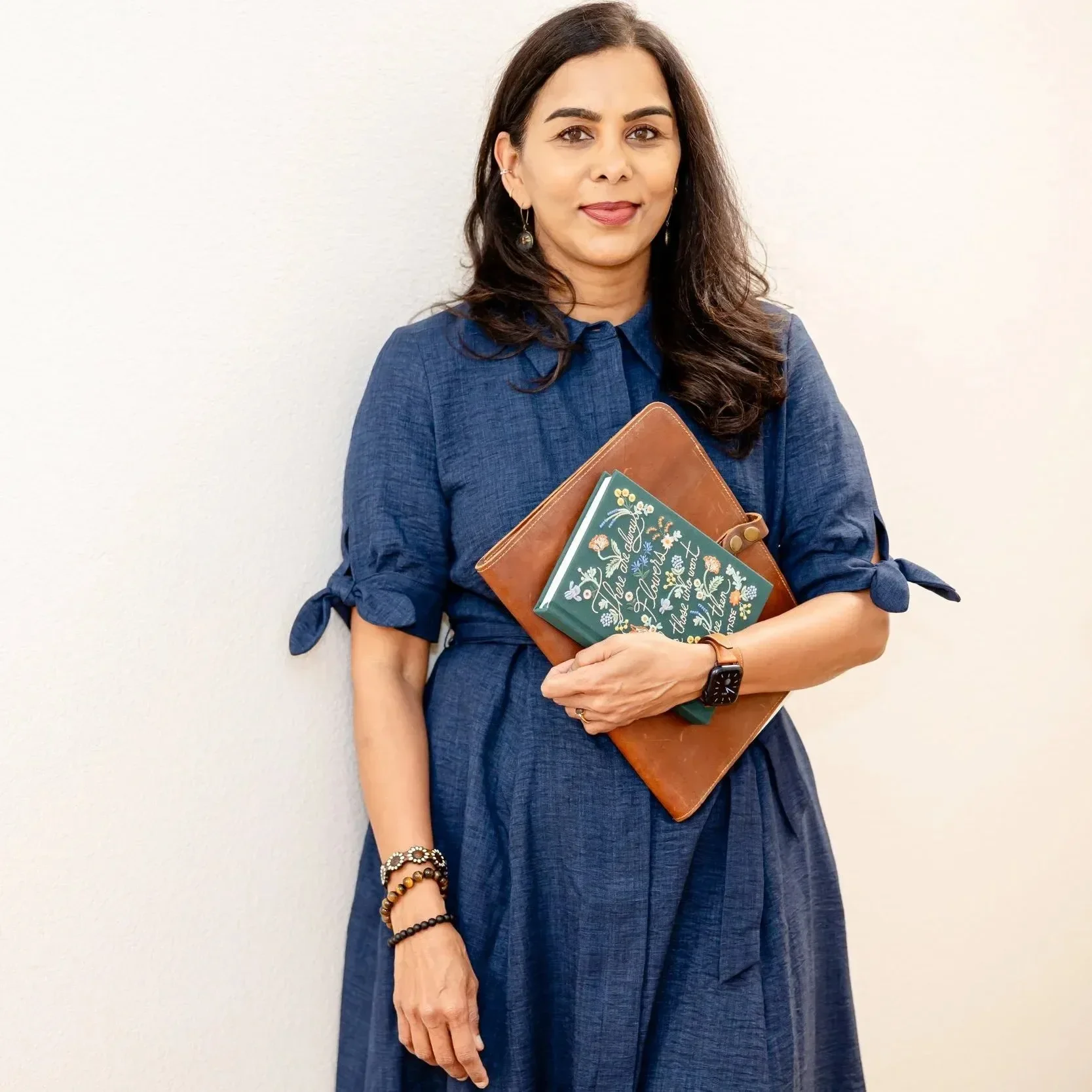 Dr. Maheswari Raja wearing a blue dress holding books, standing against a white wall, smiling slightly with her tongue slightly out.