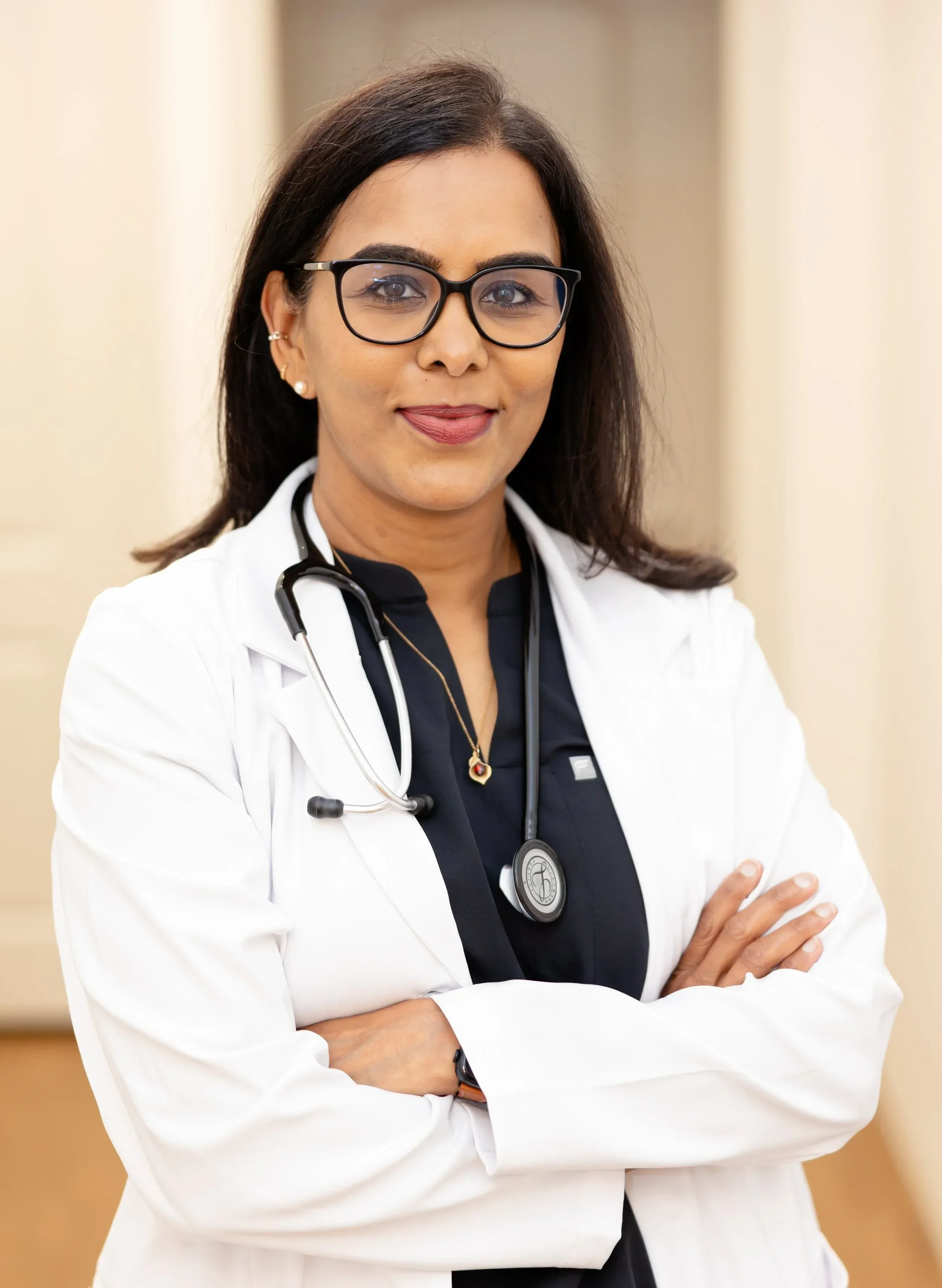 Dr. Maheswari Raja female doctor with glasses, dark hair, and pearls, wearing a white coat and stethoscope, standing with crossed arms in a medical setting.