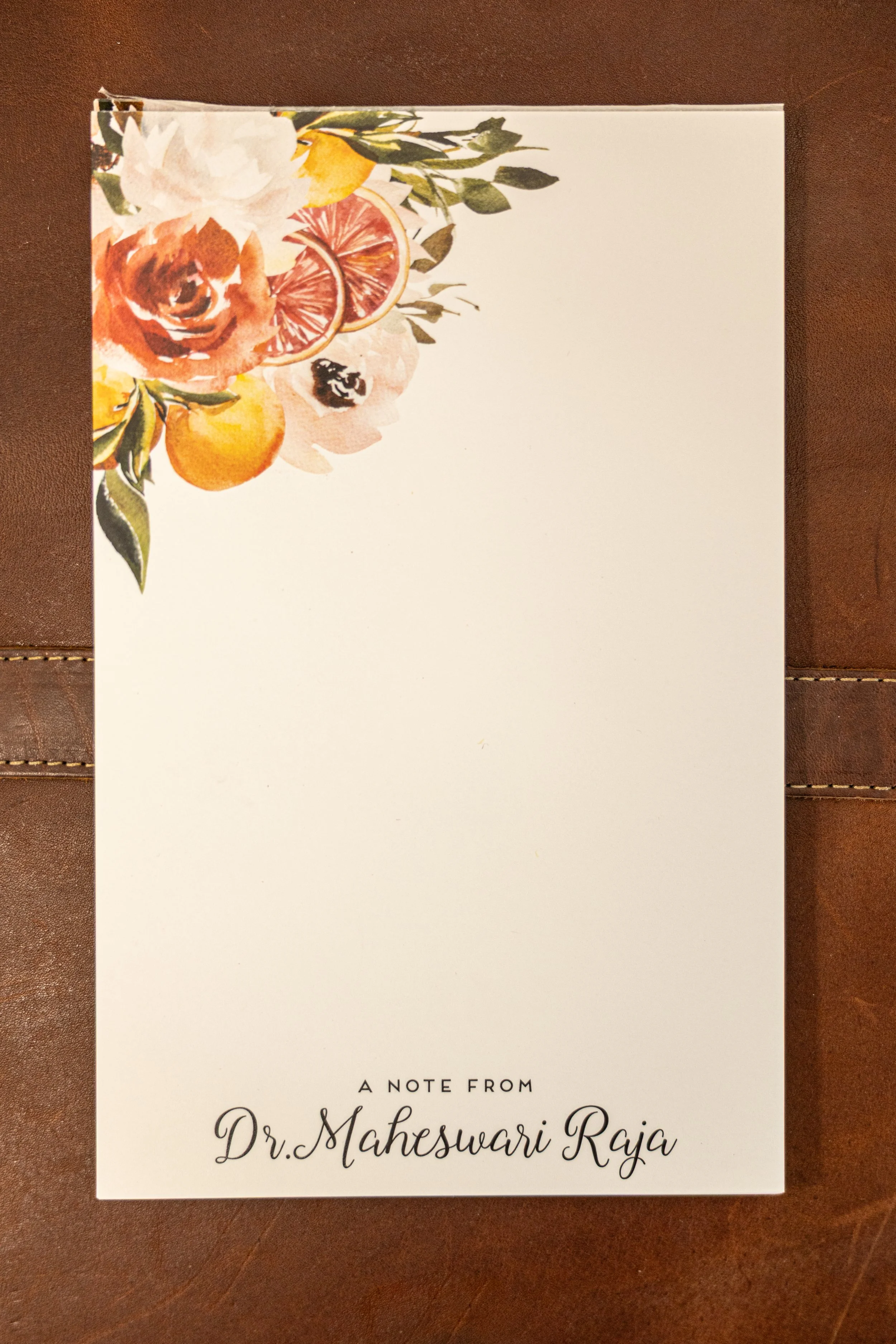 A white notepad with colorful floral and citrus fruit watercolor illustrations in the top left corner, placed on a dark brown surface. The notepad has the text 'A NOTE FROM Dr. Maheswari Raja' in elegant script.