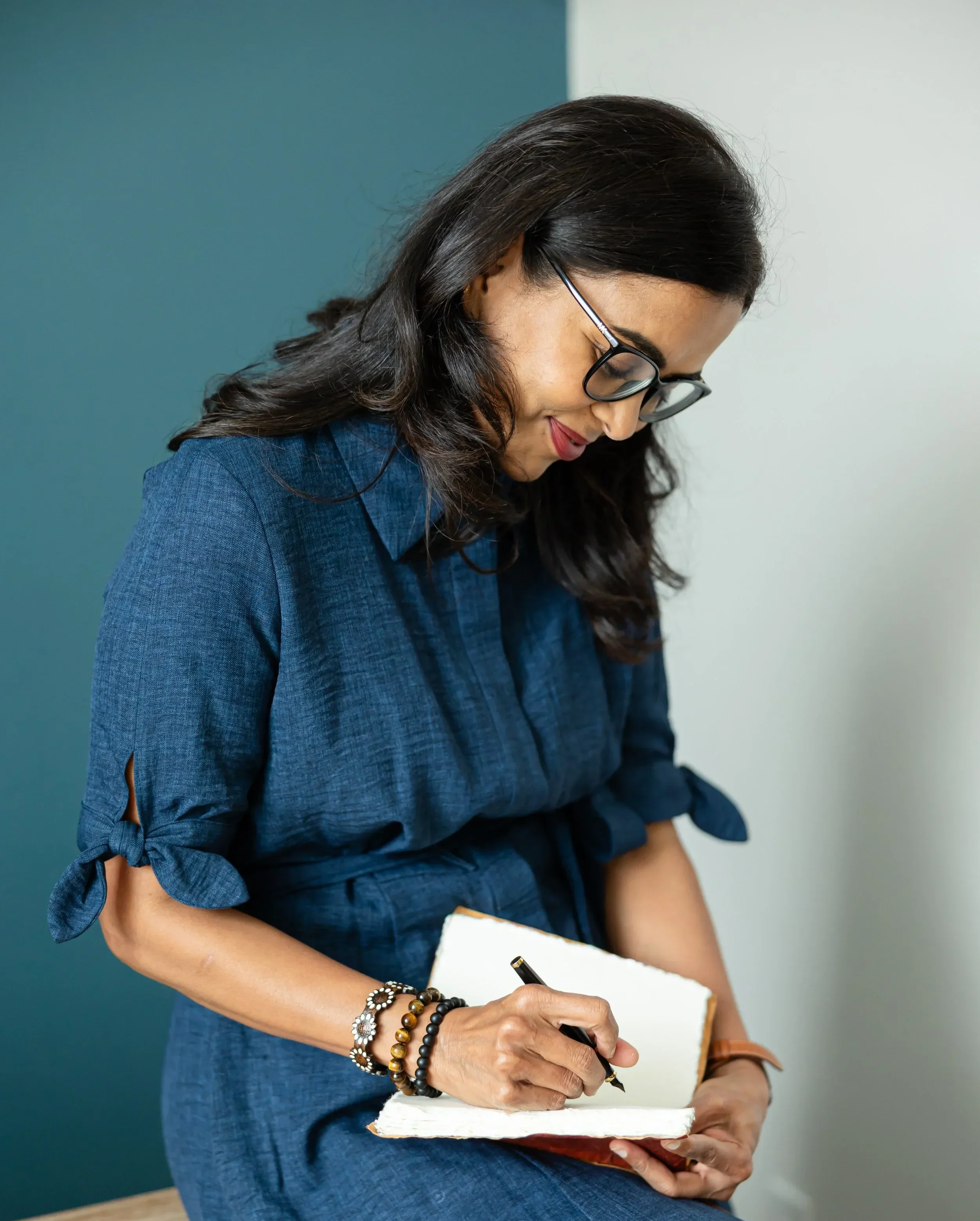 Dr. Maheswari Raja with dark wavy hair, wearing glasses and a dark blue dress, is sitting and writing in a notebook with a black pen. She is adorned with multiple bracelets on her wrist.