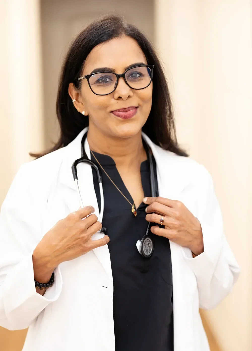 Dr. Maheswari Raja with long dark hair, wearing glasses, a white coat, and a stethoscope around her neck, standing indoors.