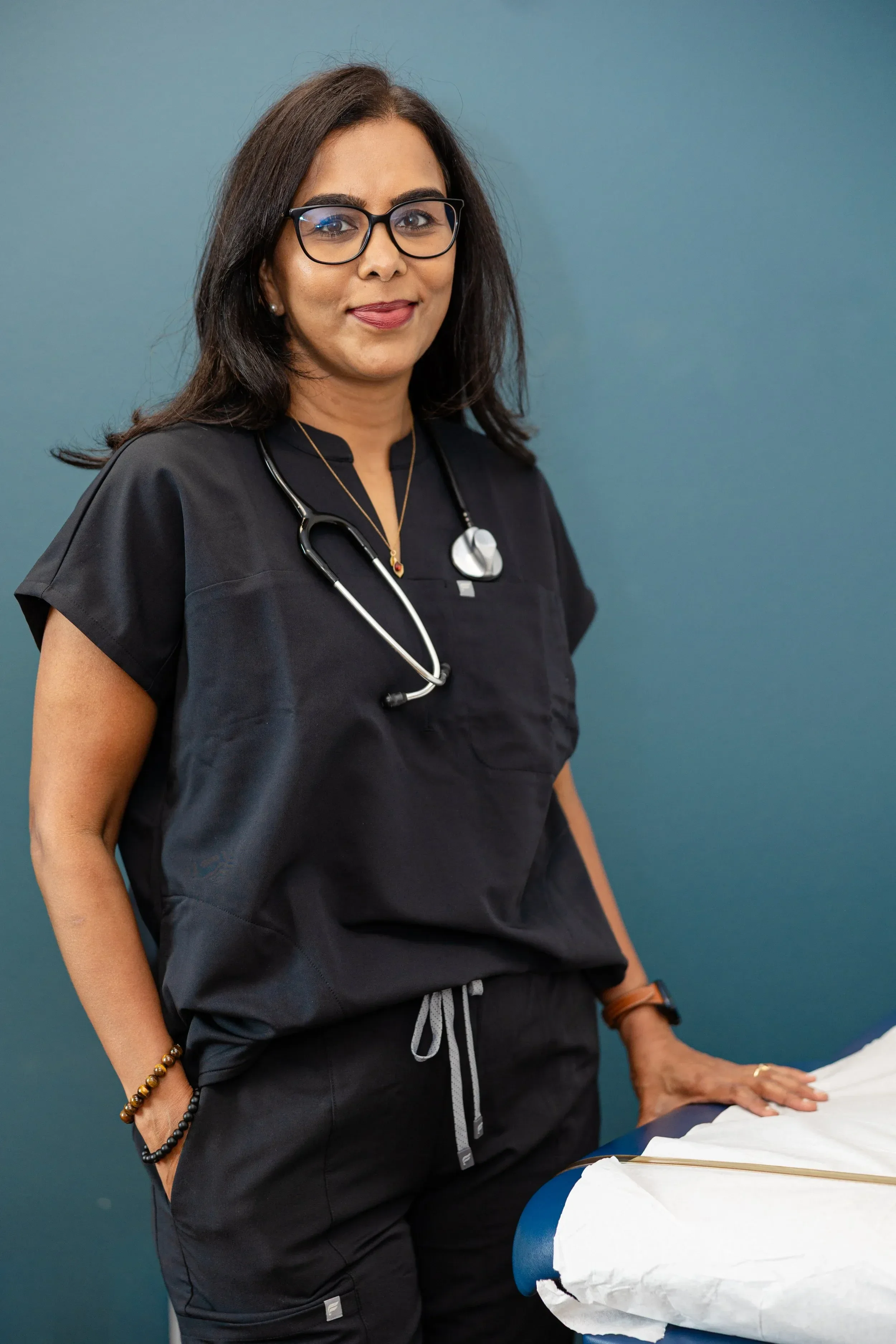 Dr. Maheswari Raja, a female healthcare professional with dark hair, glasses, wearing black scrubs, and a stethoscope around her neck, standing in front of a blue wall, with her hand resting on a medical examination table covered with white paper.