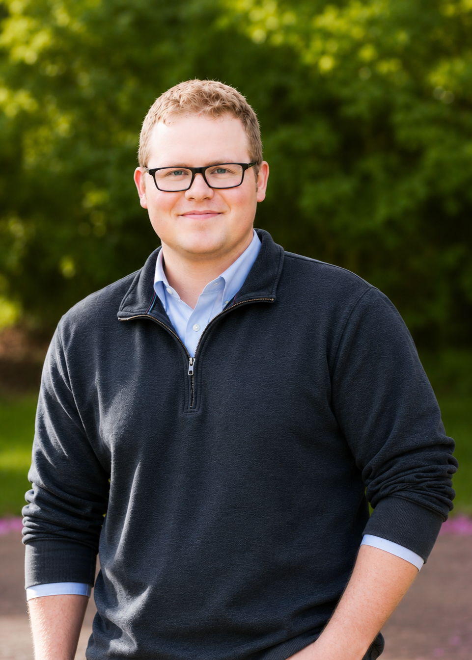 A young man with glasses and short brown hair standing outdoors with a slight smile, wearing a blue button-down shirt and a black zip-up sweater, with a green foliage background.