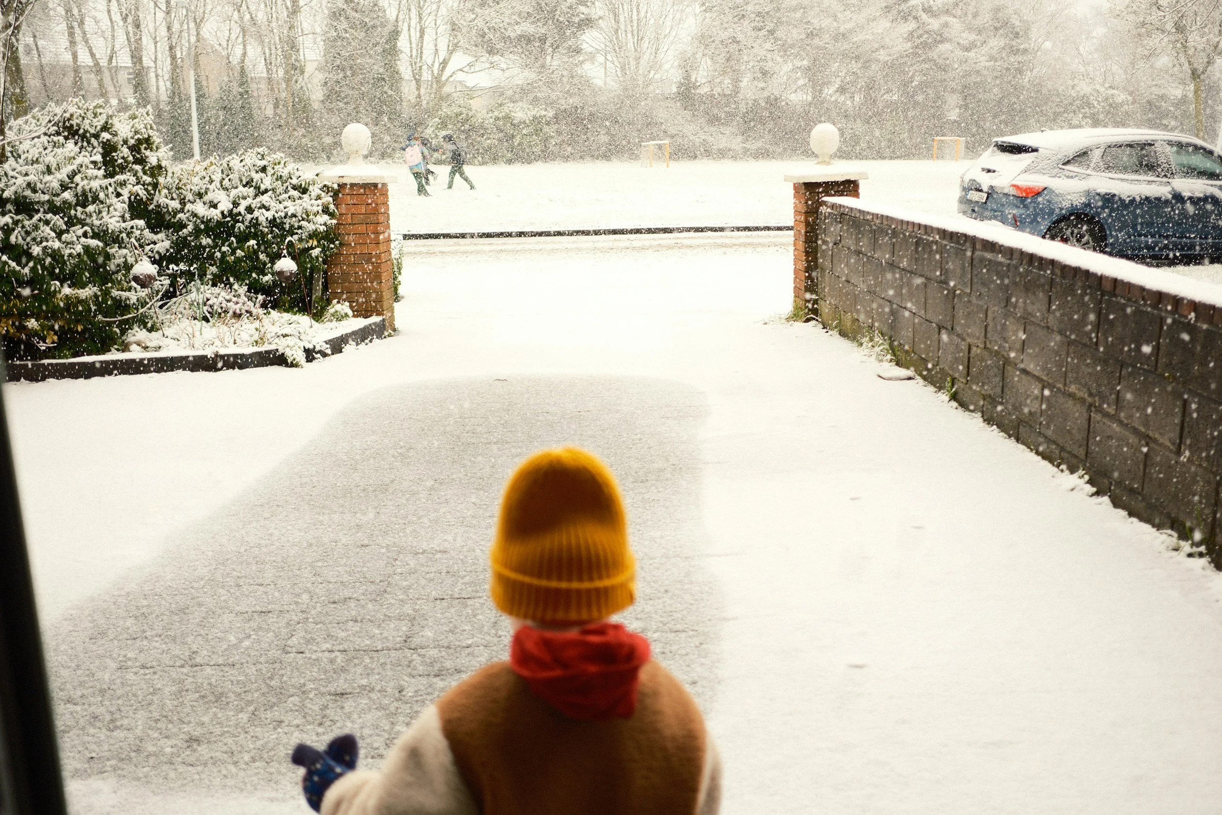 A view from inside a house showing a snow-covered front yard and street with trees, parked cars, and two children playing in the snow in the distance. A stuffed animal, resembling a dog, wearing a yellow hat and red scarf, is in the foreground.
