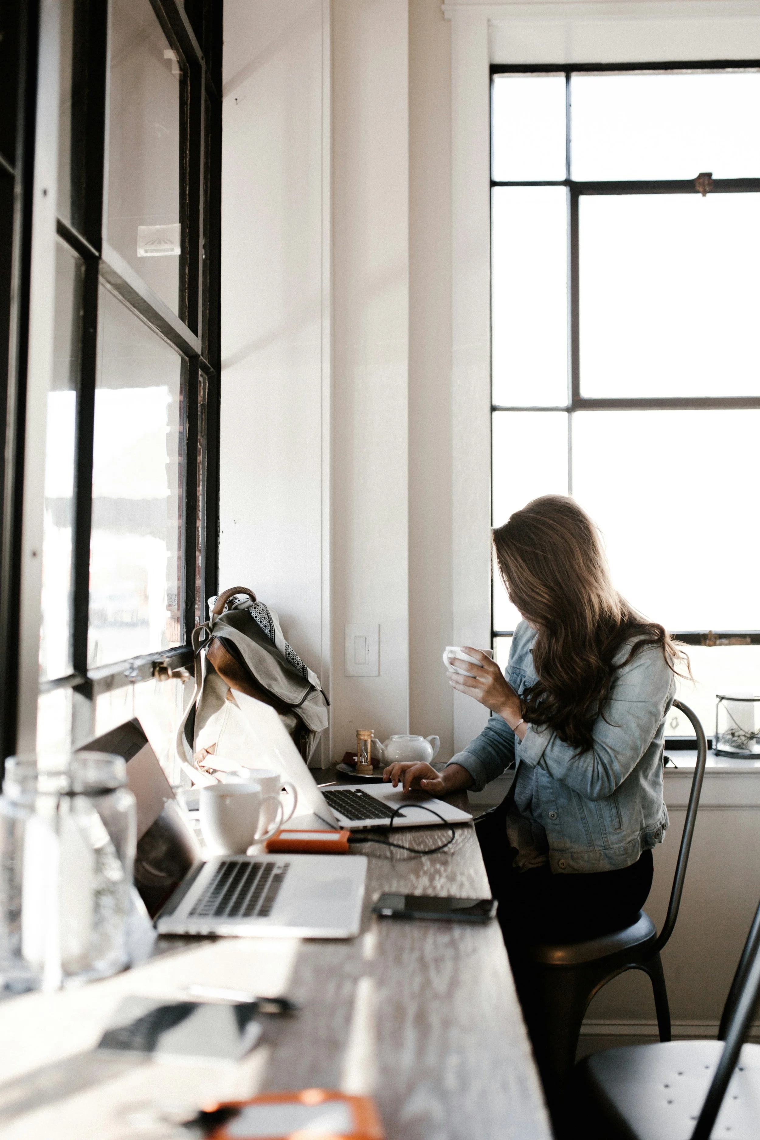 A woman with long brown hair, wearing a denim jacket, sitting at a cluttered wooden table with a laptop, smartphone, and notebooks, in a bright room with large windows and black metal frames, engaging with her phone.