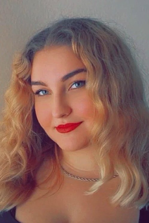 Close-up photo of a woman with curly blonde hair, blue eyes, red lipstick, and wearing a silver necklace.