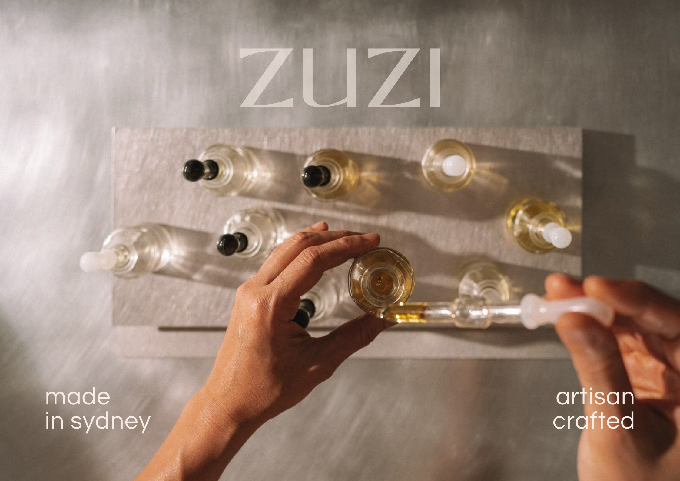 A person holding a small clear bottle with golden liquid. There are several similar bottles on a gray surface with the words "ZUZI made in sydney artisan crafted" overlaid on the image.