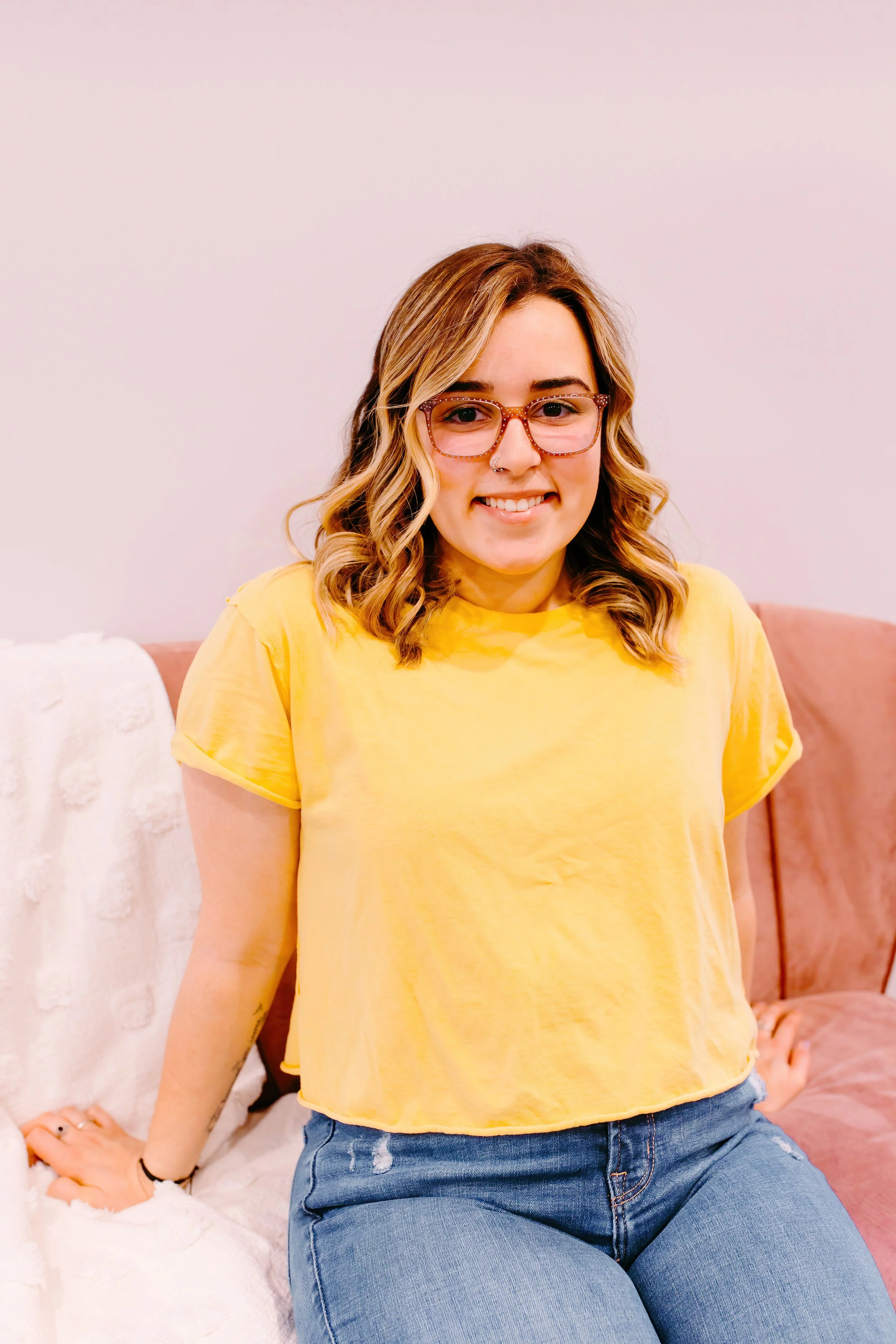 hairstylist with brown hair, highlights in her hair and glasses, sitting relaxed on a couch