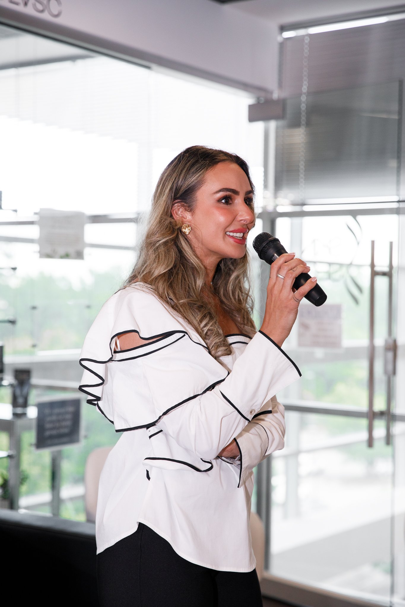 A woman with long wavy blonde hair, wearing a white blouse with black trim and black pants, holding a microphone and speaking indoors with bright windows in the background.