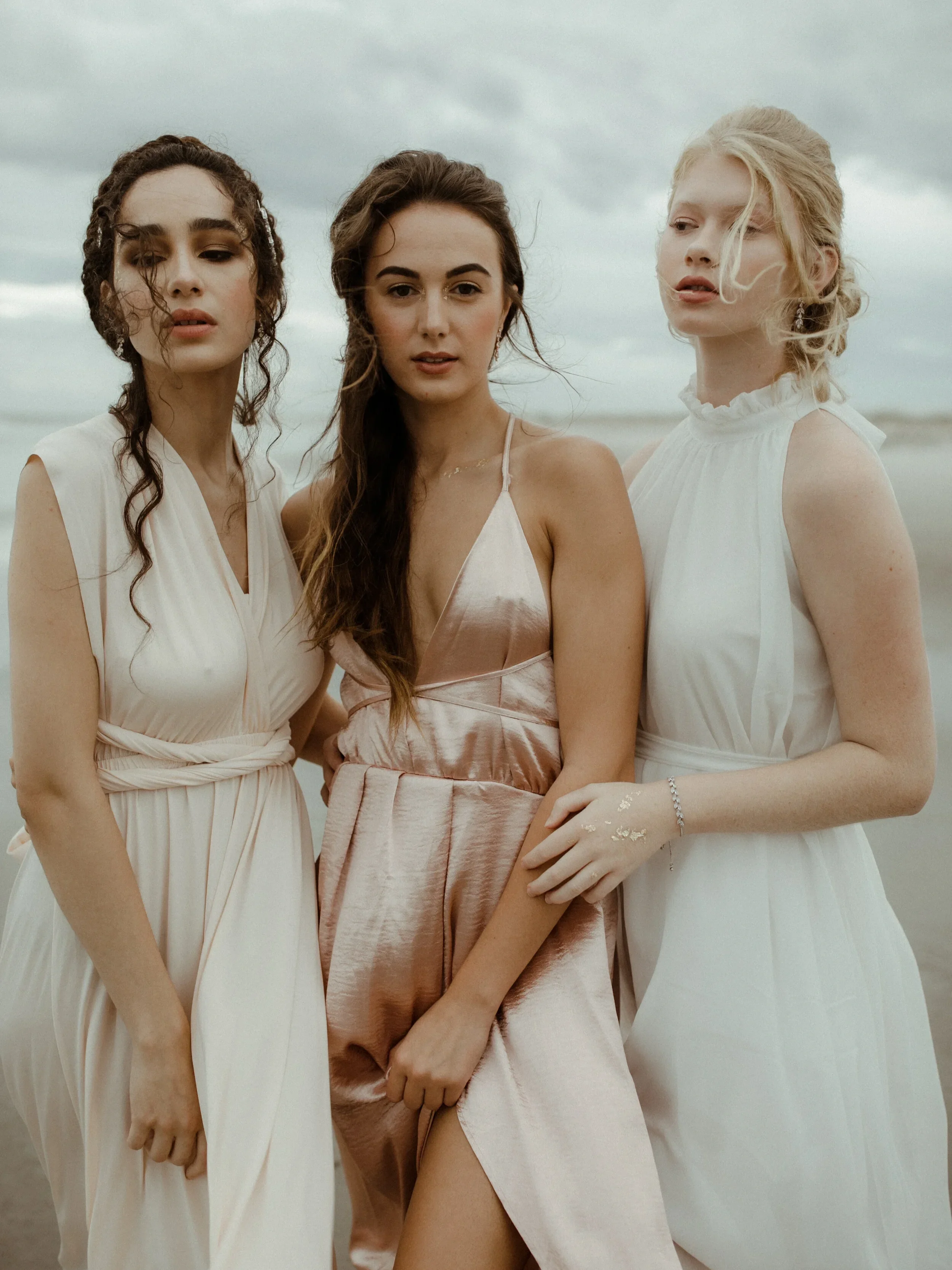 Three women standing close together on a cloudy day, wearing light-colored dresses, with a beach or ocean background.