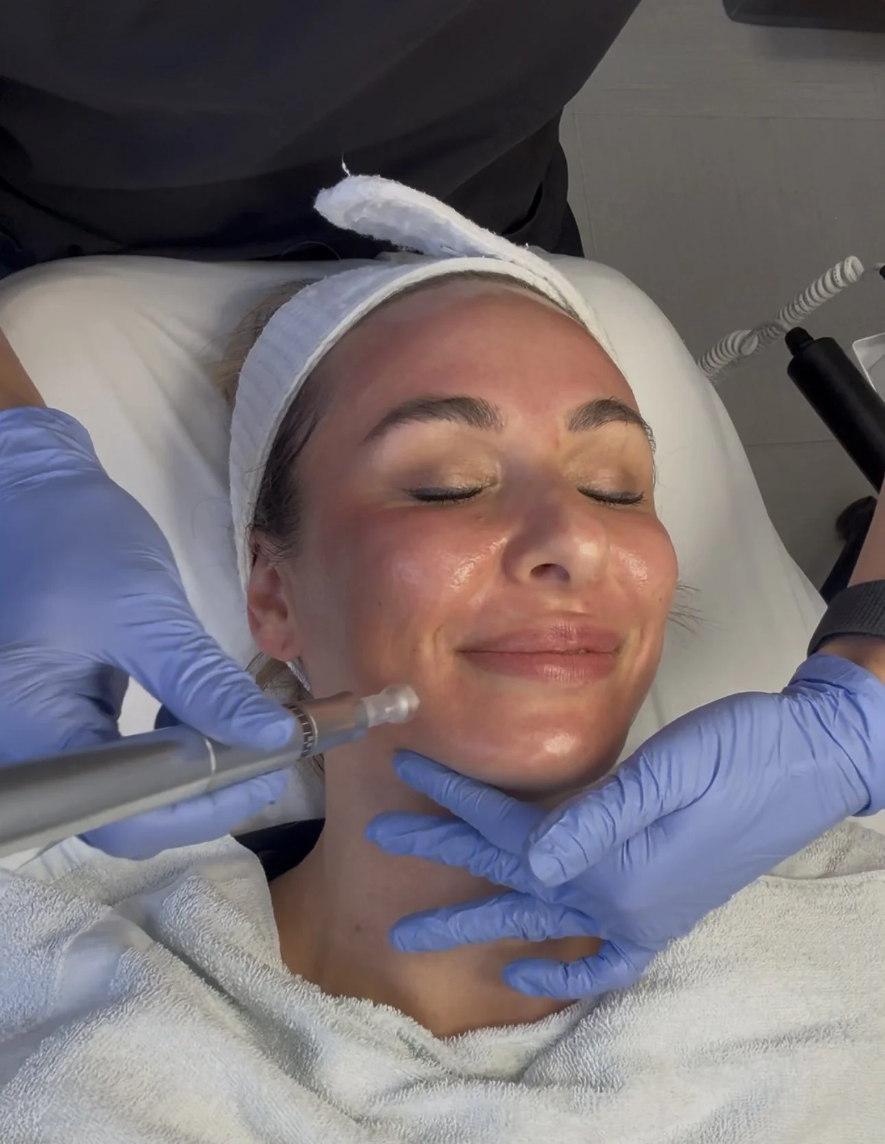 A woman receiving a facial treatment in a spa or medical clinic, with a relaxed and content expression, a headband, and a towel wrapped around her shoulders, while a professional wearing blue gloves administers a treatment using a device.