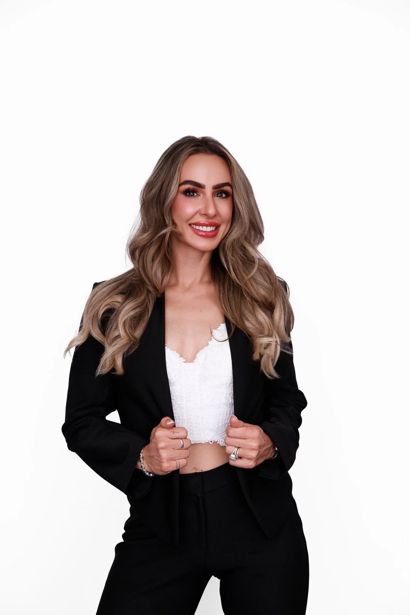 A smiling woman with long, wavy blonde hair, wearing a black blazer and pants, a white lace top, and rings on her fingers, standing against a plain white background.