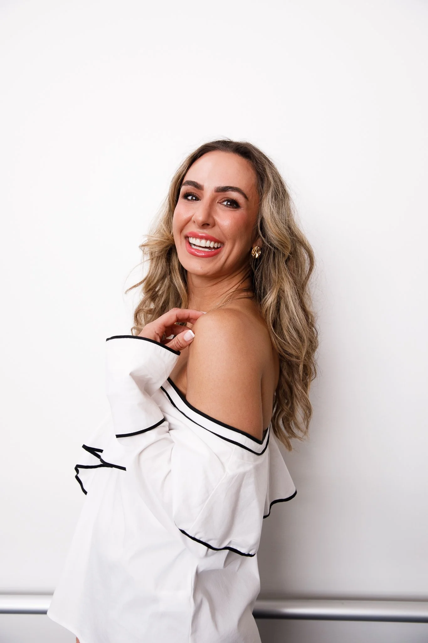 A young woman with long wavy blonde hair, smiling and showing her teeth, is posing against a plain white background. She is wearing a white off-the-shoulder top with black trim and gold hoop earrings.