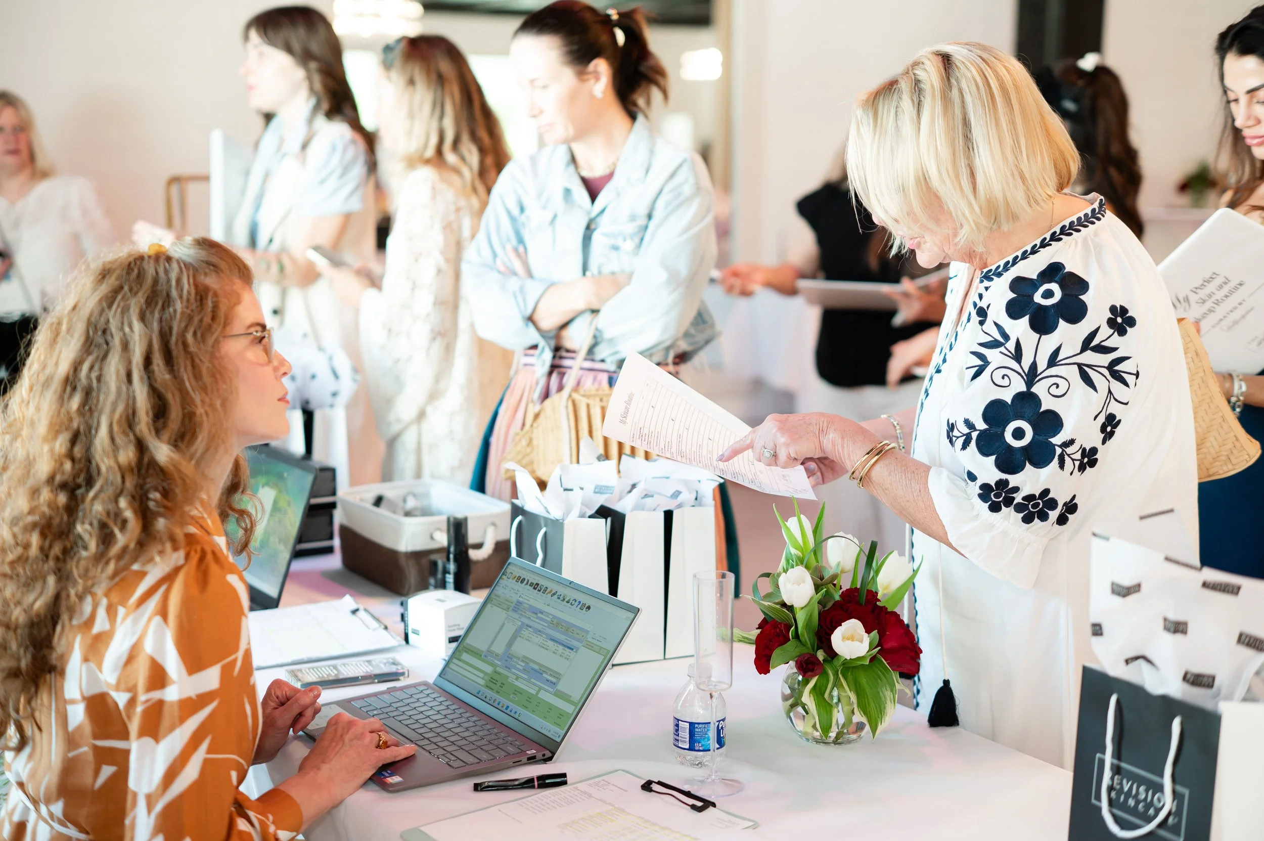 Branding or registration table at an event with women interacting, some looking at paperwork and laptops, a woman with blonde hair in a white and blue floral dress reading a paper, a woman with curly hair seated at the computer, flowers, and various event materials on the table.