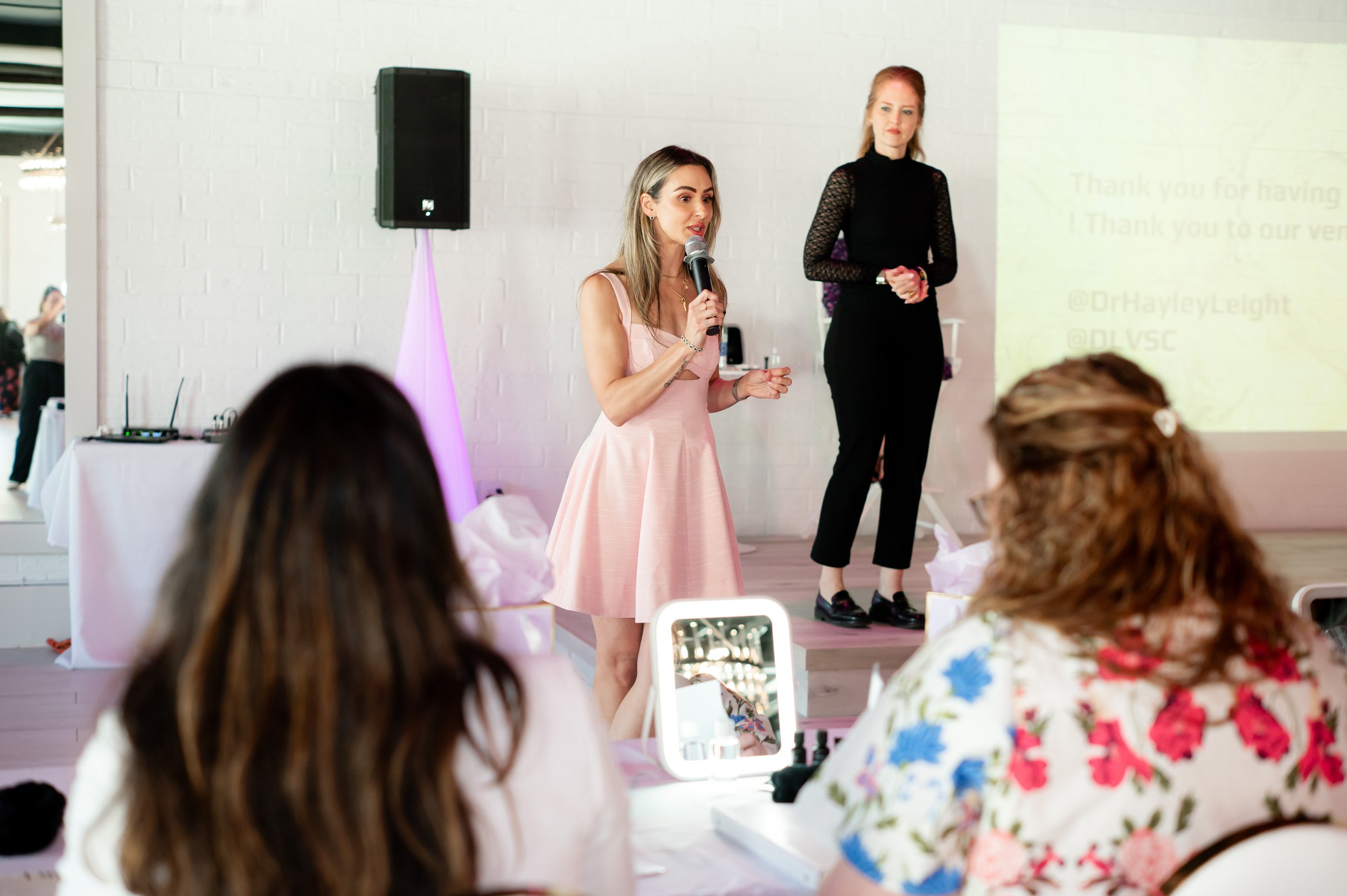 A woman in a pink dress is speaking into a microphone at an indoor event, with another woman standing behind her. The audience is seated, and some are taking photos.
