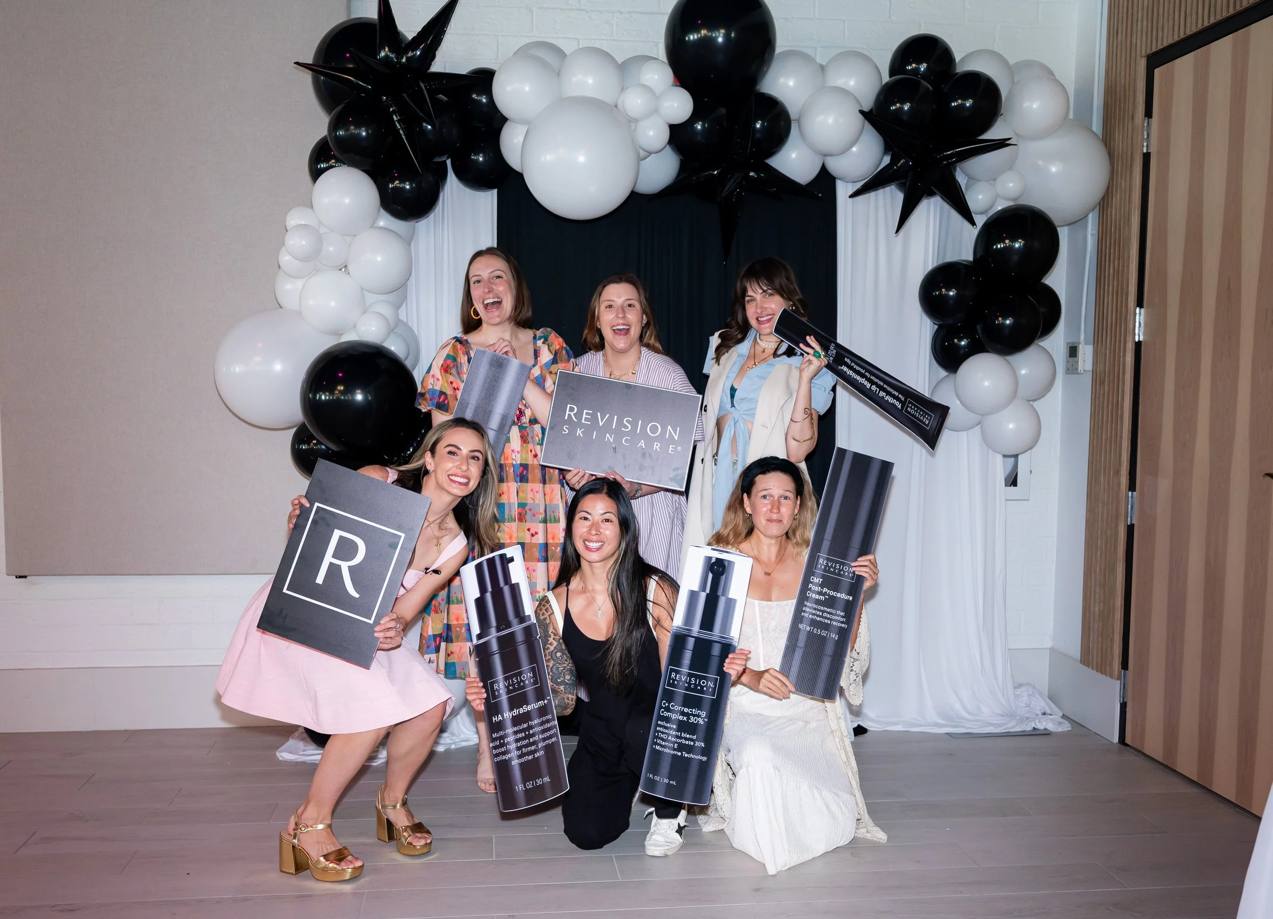 A group of women smiling and holding skincare product posters at an event, standing in front of a balloon arch with black, white, and star-shaped balloons.