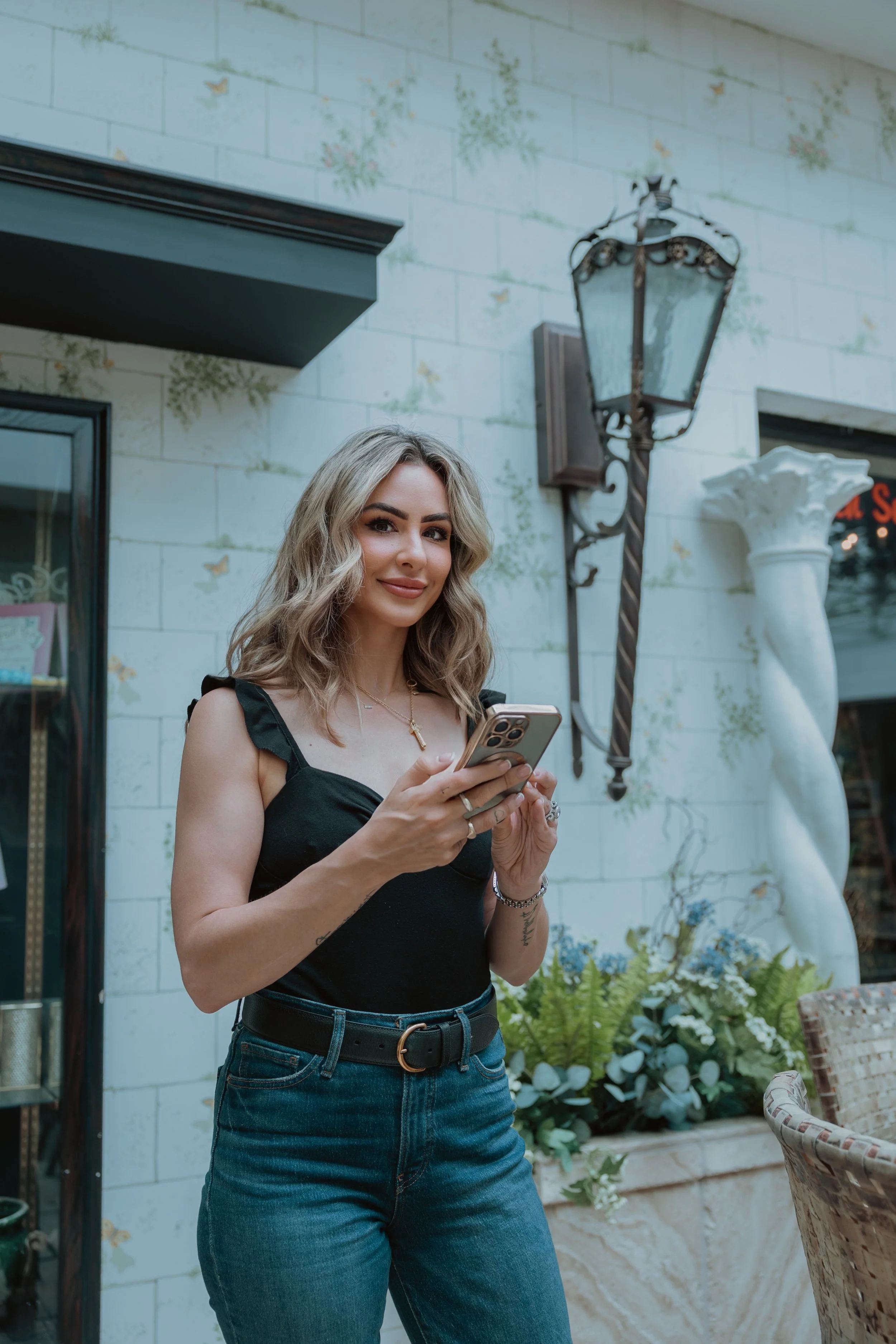 A woman with blonde, wavy hair and wearing a black sleeveless top, blue jeans, and a belt, stands outside holding a smartphone and smiling.