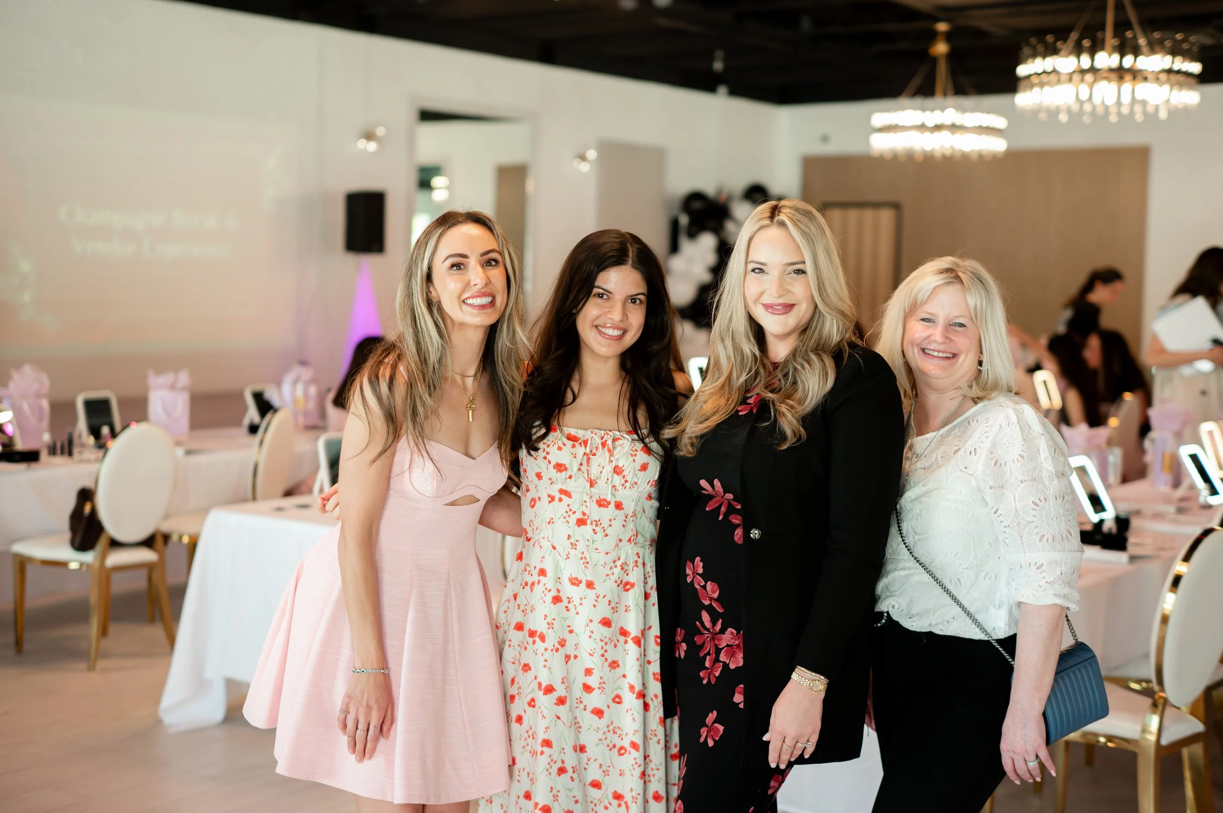 Four women smiling and posing together indoors at a formal event with decorated tables.