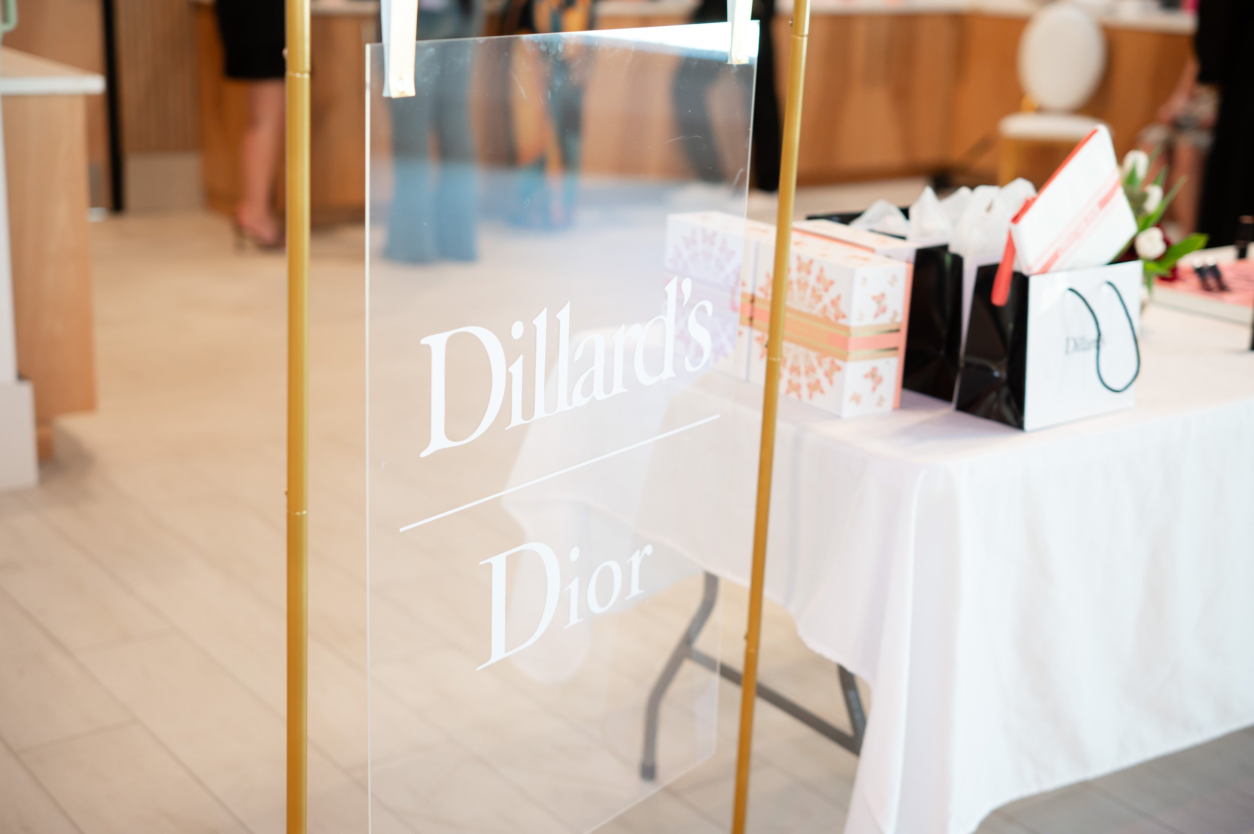 A clear acrylic sign with white text that reads "Dillard's Dior" positioned on a gold-colored stand at an event or store display, with a table in the background holding gift bags and wrapped boxes.