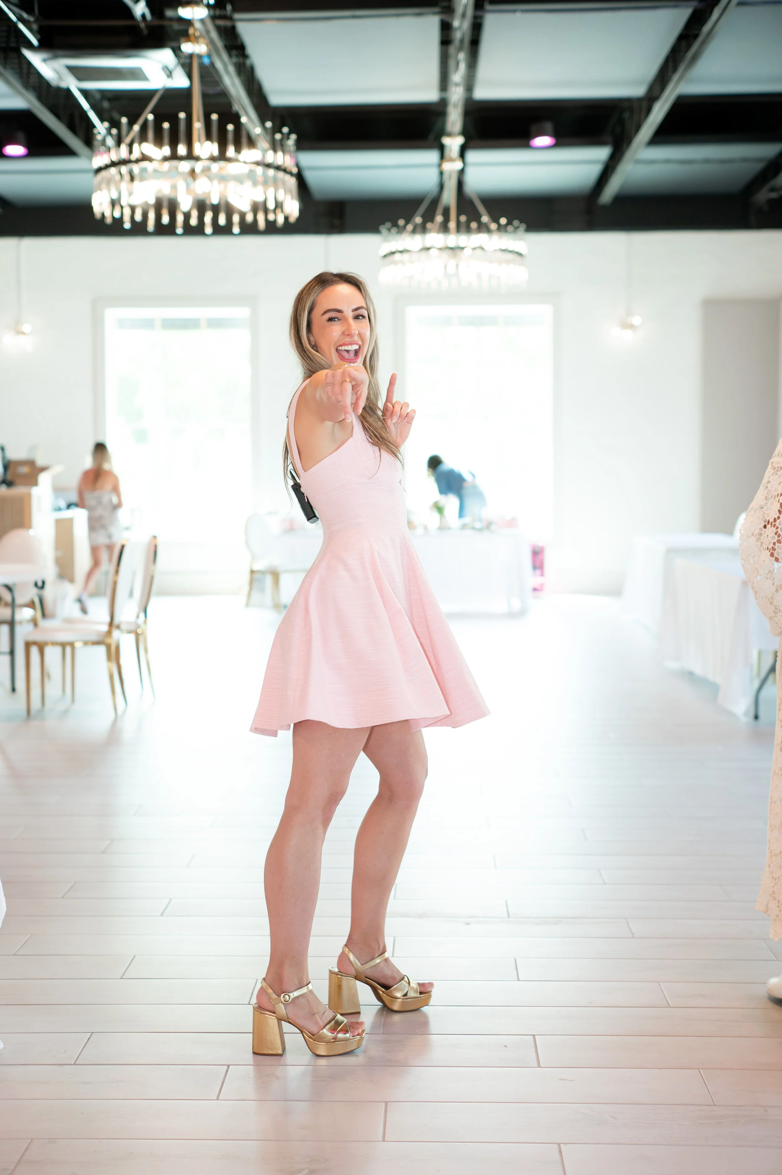 A woman in a pink dress, wearing gold high-heeled shoes, is smiling and pointing towards the camera in a brightly lit room with chandeliers and large windows.