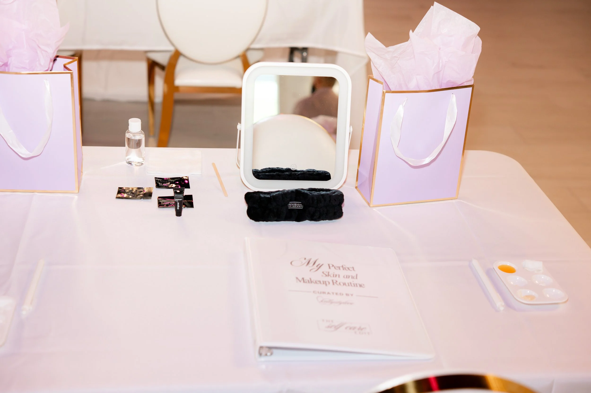 Display table with skincare and makeup products, including a folded black headband, small sample packets, a mirror, a small clear bottle of liquid, and a sign reading 'My Perfect Skin and Makeup Routine'.