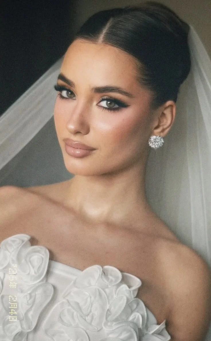 Close-up of a woman with dark hair styled in an elegant updo, wearing natural makeup with defined eyes, and large sparkling earrings. She is dressed in a white strapless dress with ruffled fabric details and has a sheer veil in the background.