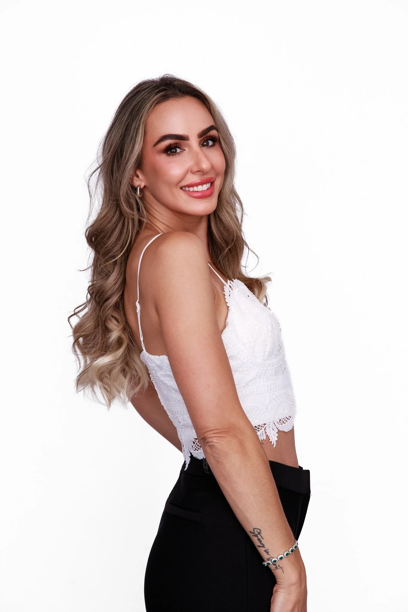 A young woman with long wavy blonde hair, wearing a white lace crop top and black pants, smiling at the camera against a plain white background.
