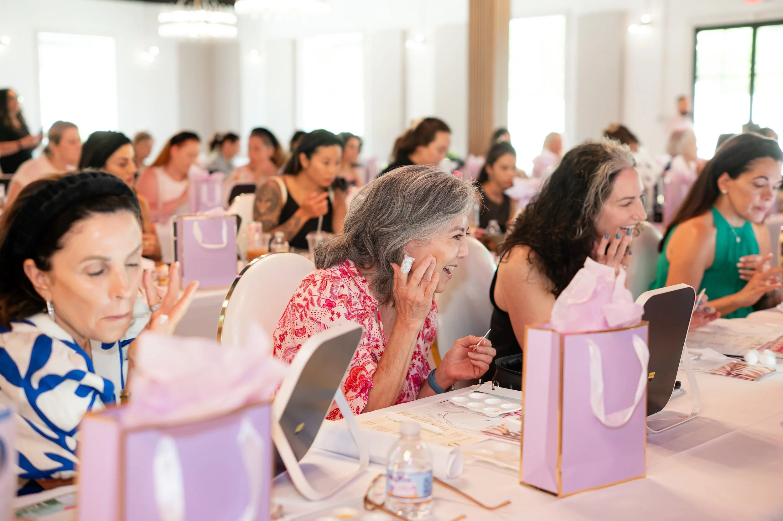 A group of women in a bright, well-lit room, sitting at tables with pink gift bags, engaged in a joyful activity. Some women are smiling and laughing, with a few holding beauty or skincare products.