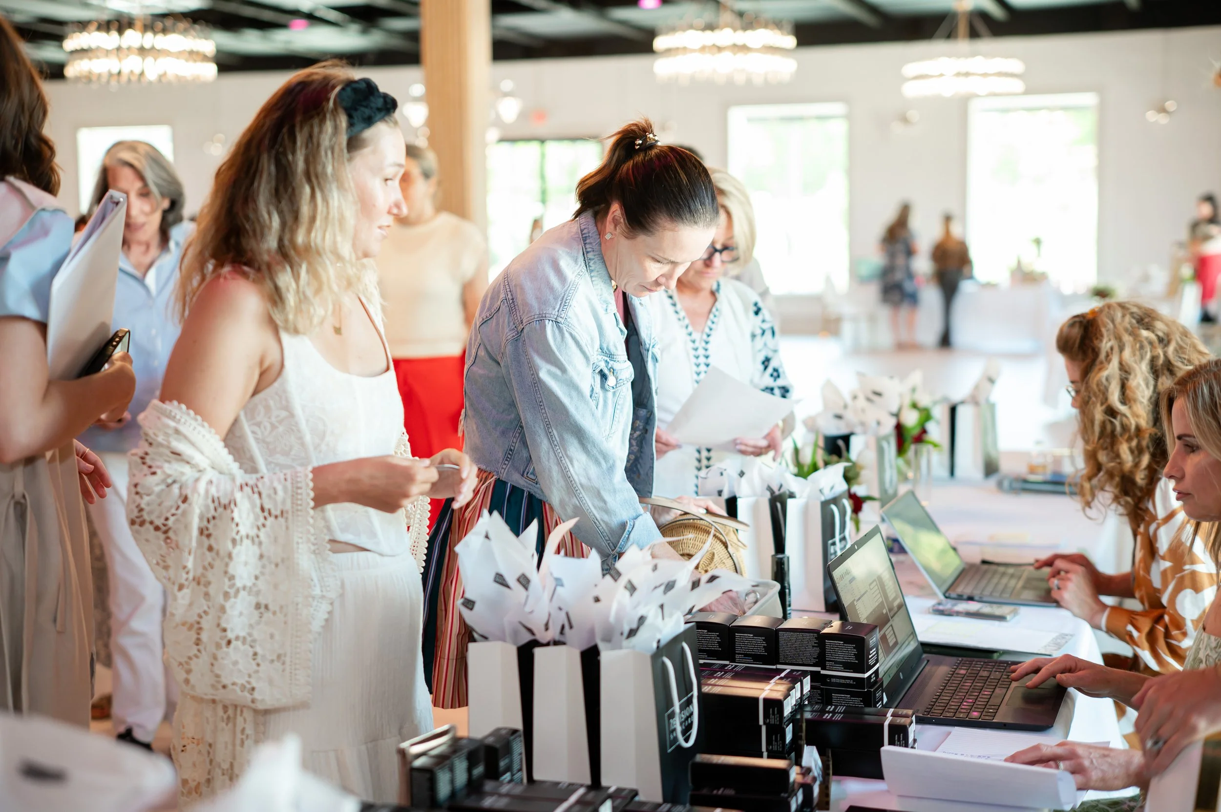 People shopping at a booth with laptops and gift bags in a brightly lit indoor market or event.