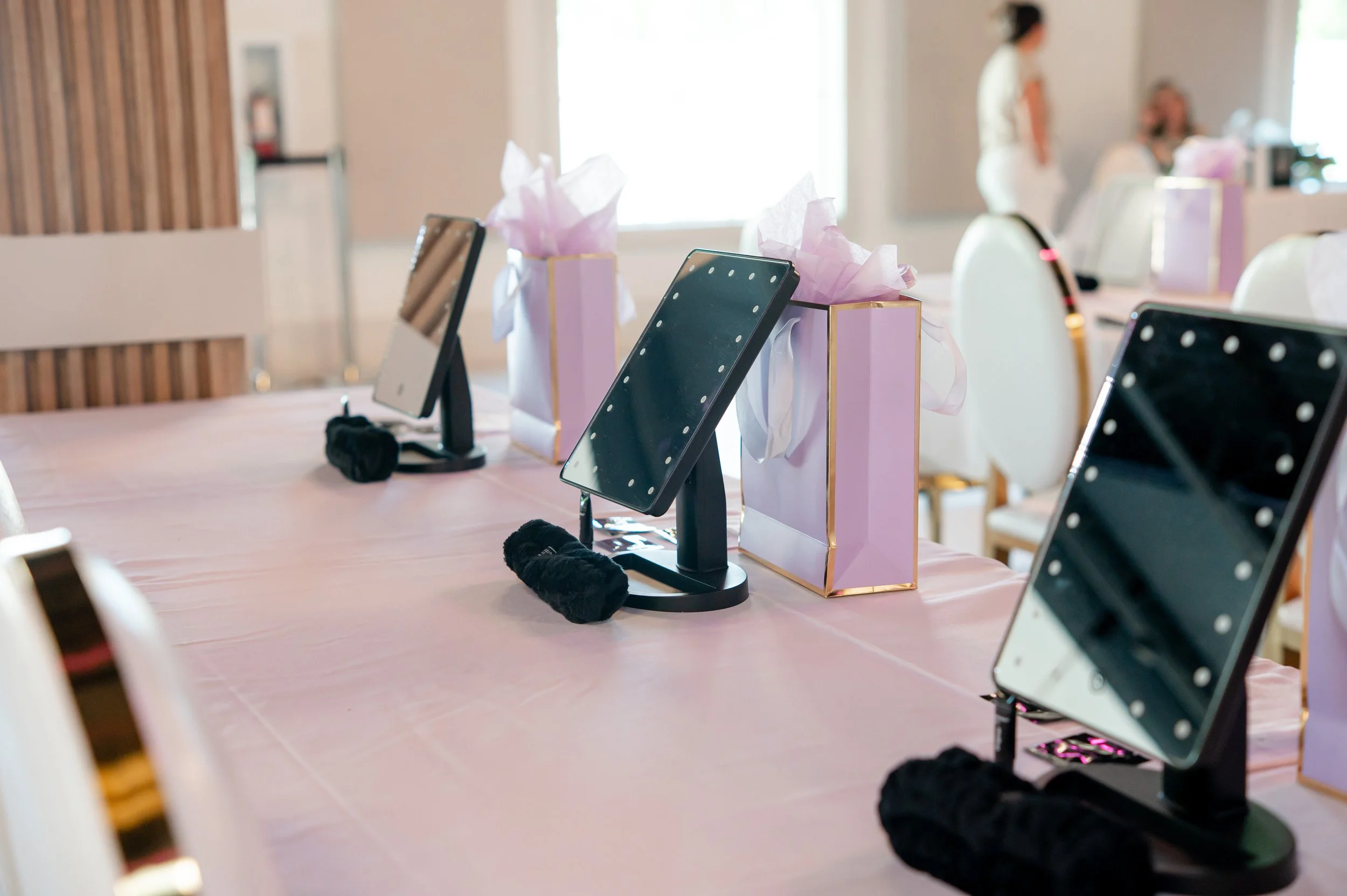 Table decorated with pink gift boxes with tissue paper, each featuring a small mirror or photo frame on a stand, in a room with chairs and people in the background, likely set up for a celebration or event.