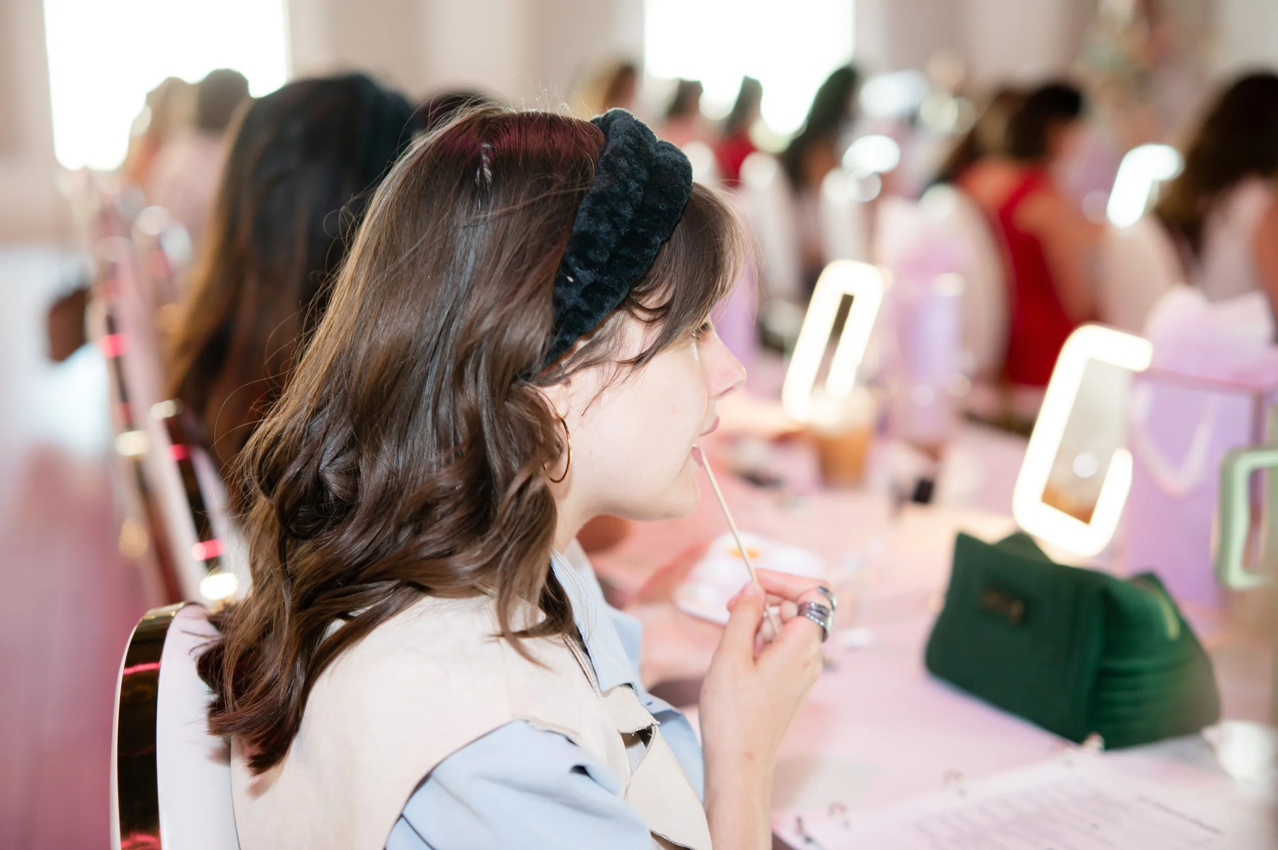 A woman with wavy brown hair wearing a black headband is sitting at a makeup or beauty counter, holding a small object in her hand, with other women sitting at similar stations in the background.