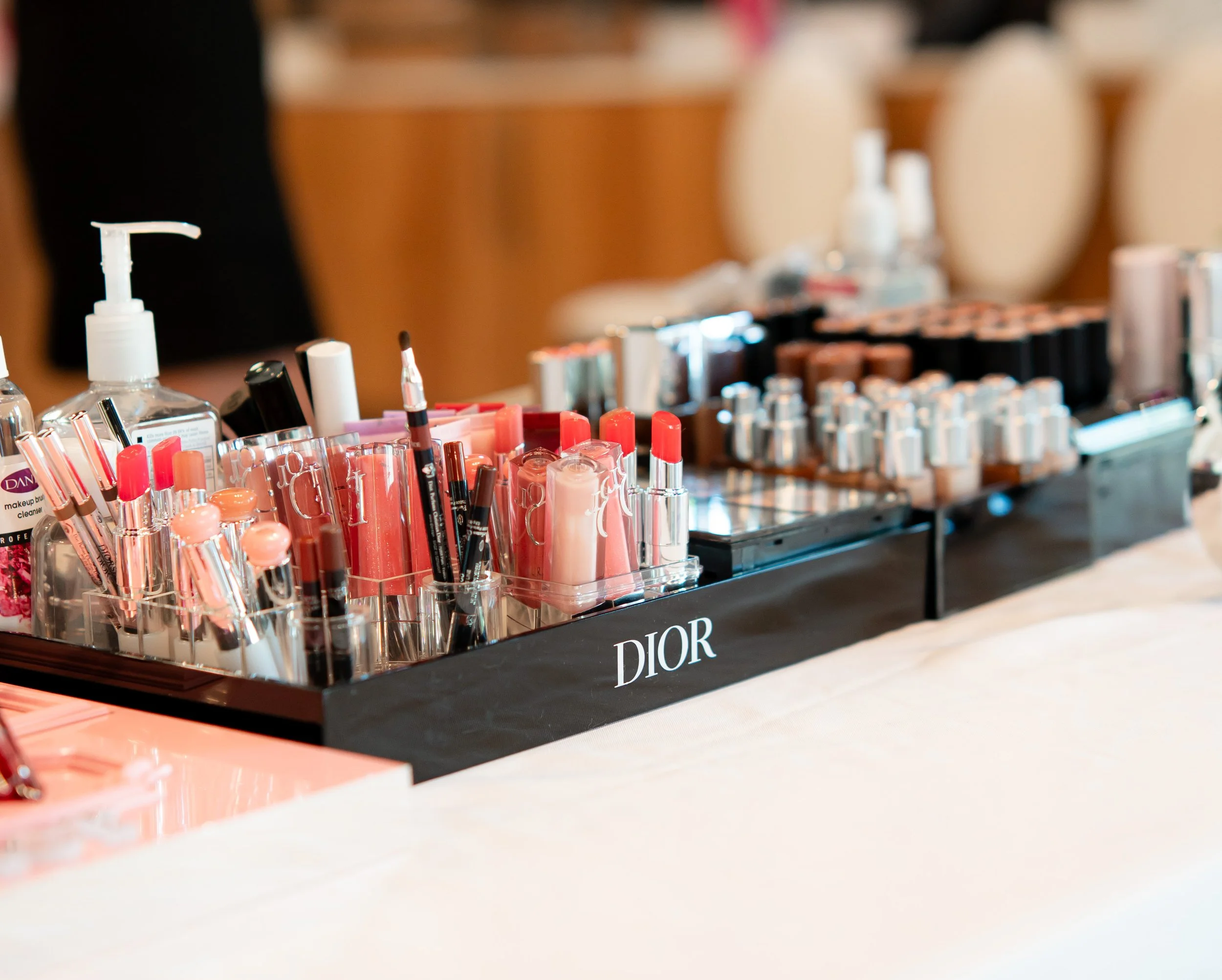 A display of various lipsticks and makeup products arranged on a black Dior-branded tray with a background of blurred chairs.