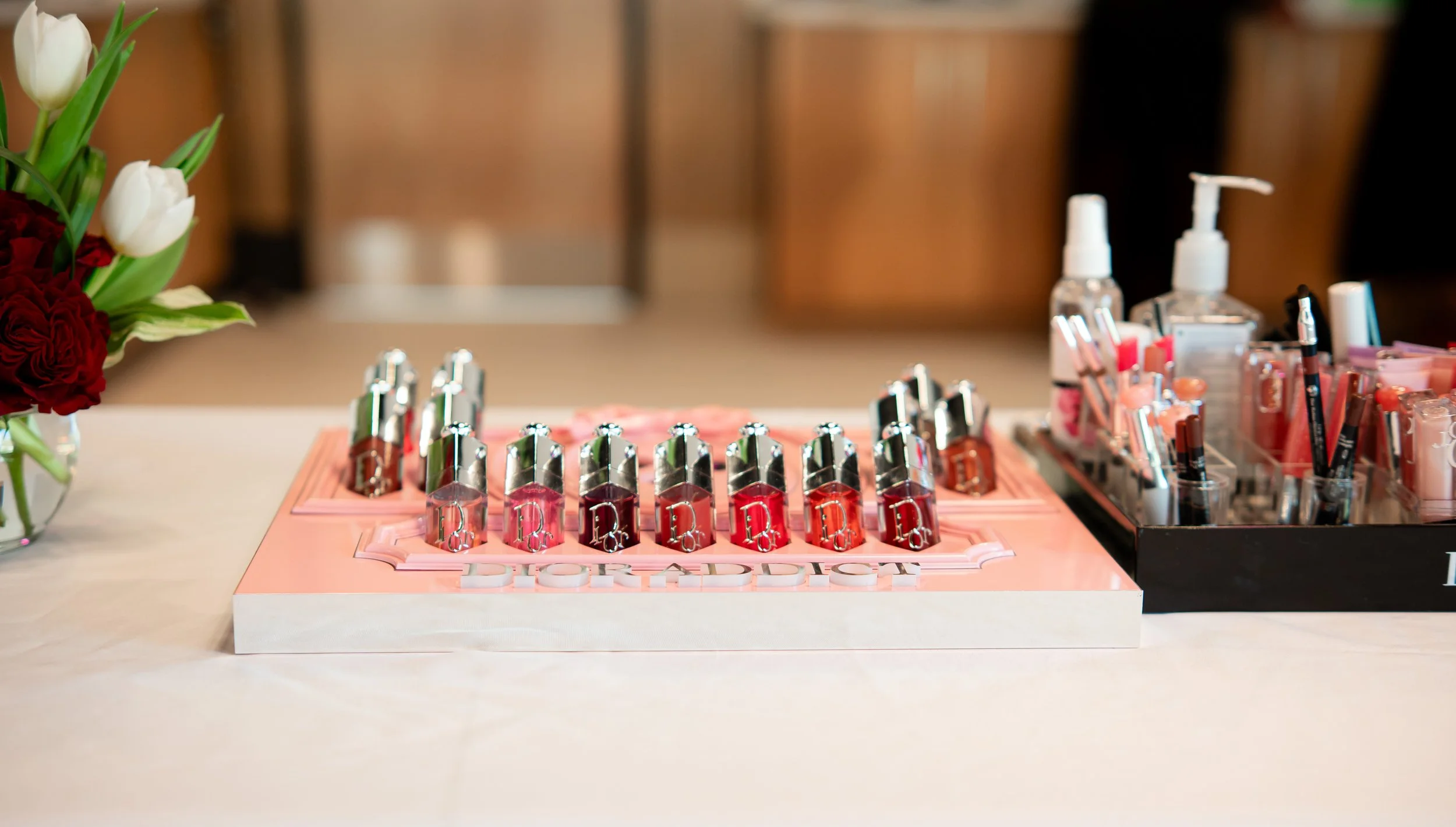 A display of various lipsticks and lip glosses, arranged on pink and black trays, with a bouquet of white and red flowers on the side.